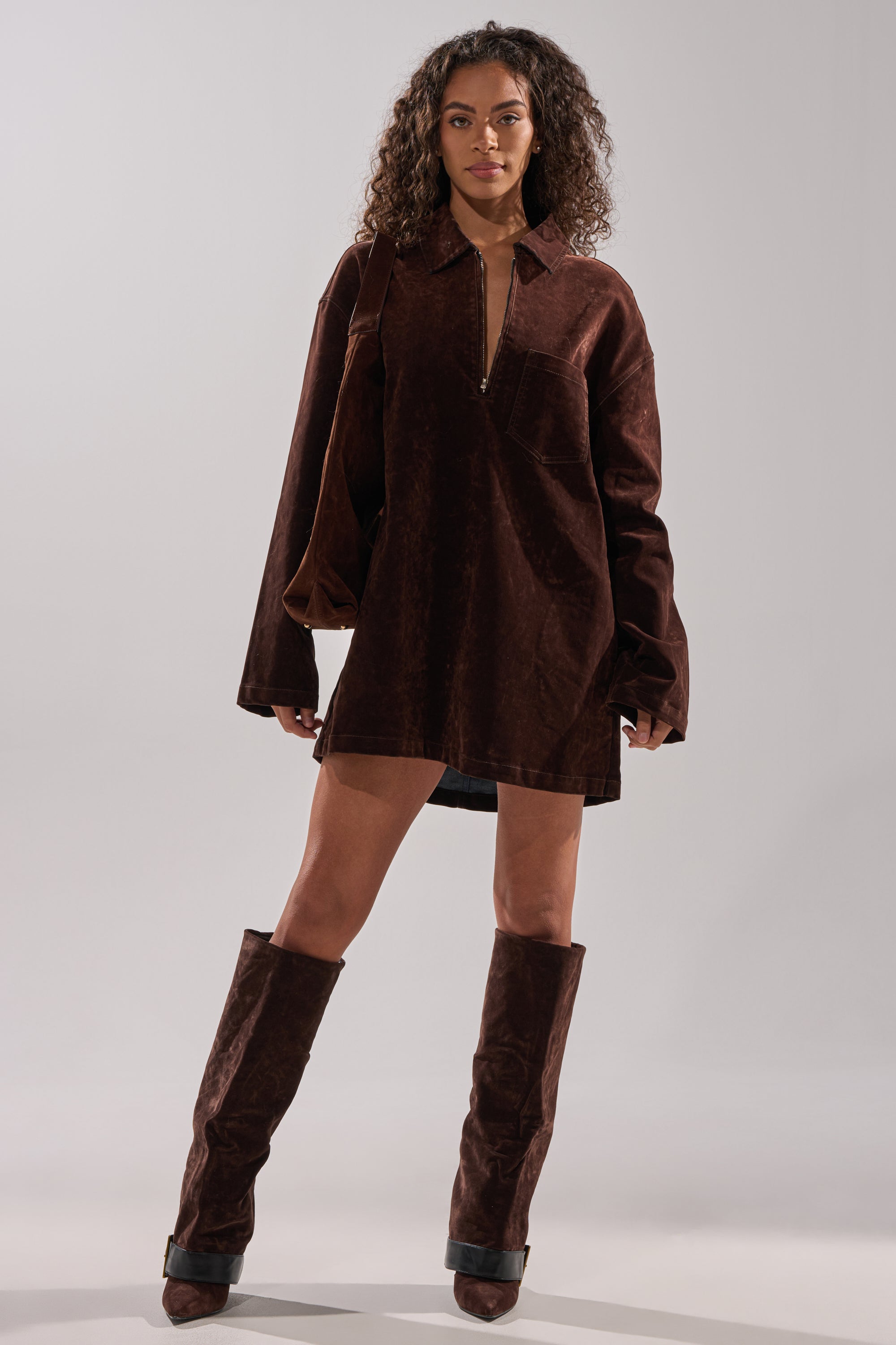 A woman with curly hair stands confidently in the COOL GIRL OVERSIZED VELVET DENIM SHIRT DRESS in chocolate brown, paired with matching knee-high boots against a plain light gray background.