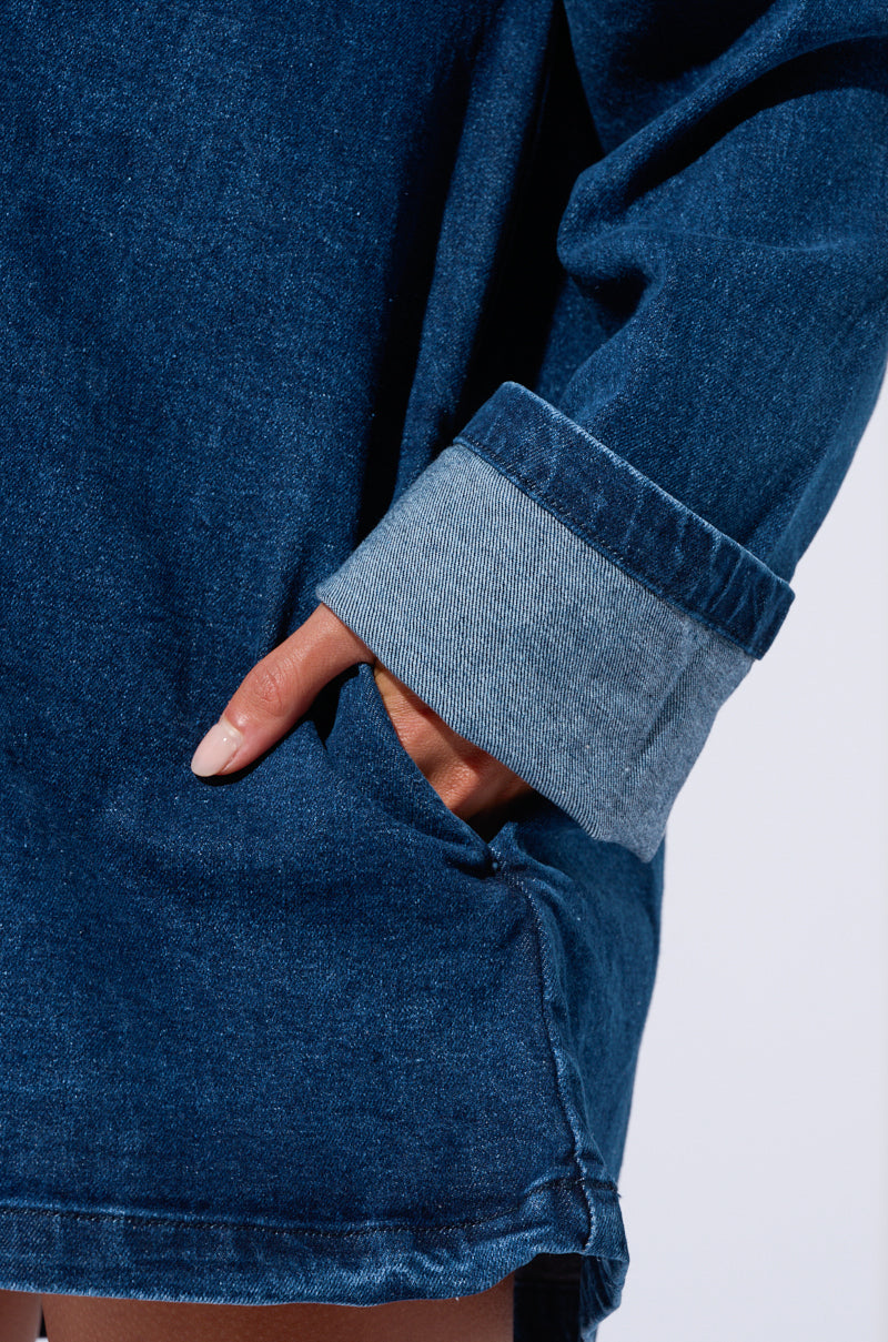 A person wearing the COOL GIRL OVERSIZED DENIM SHIRT DRESS with the sleeves partially rolled up has their hand in the dress pocket. The image is focused on the lower section of this casual-cute piece and the hand.