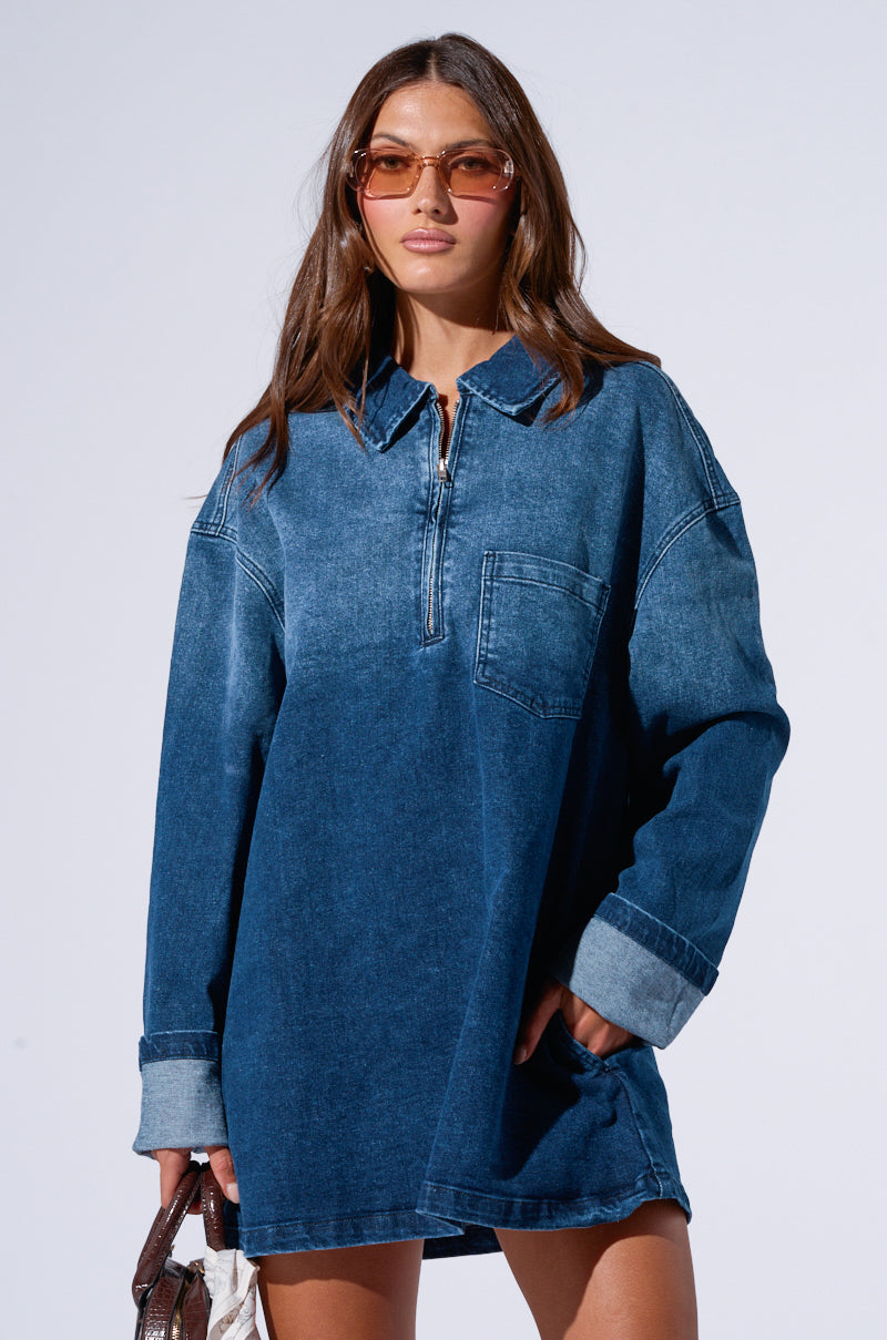 A woman with long brown hair wears the COOL GIRL OVERSIZED DENIM SHIRT DRESS made of dark wash denim, featuring rolled-up sleeves and a chest pocket. She sports oversized sunglasses and holds a handbag against a plain white background.