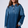 A woman with long brown hair wears the COOL GIRL OVERSIZED DENIM SHIRT DRESS made of dark wash denim, featuring rolled-up sleeves and a chest pocket. She sports oversized sunglasses and holds a handbag against a plain white background.