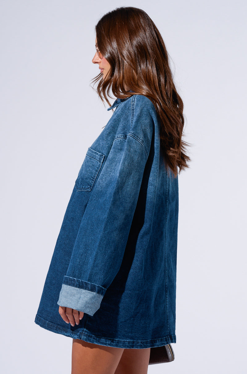 A woman with long brown hair stands in profile against a plain background, wearing the COOL GIRL OVERSIZED DENIM SHIRT DRESS. The dark wash piece, complete with rolled-up sleeves and a chest pocket, gives her look a relaxed yet stylish vibe as she appears to be looking forward.