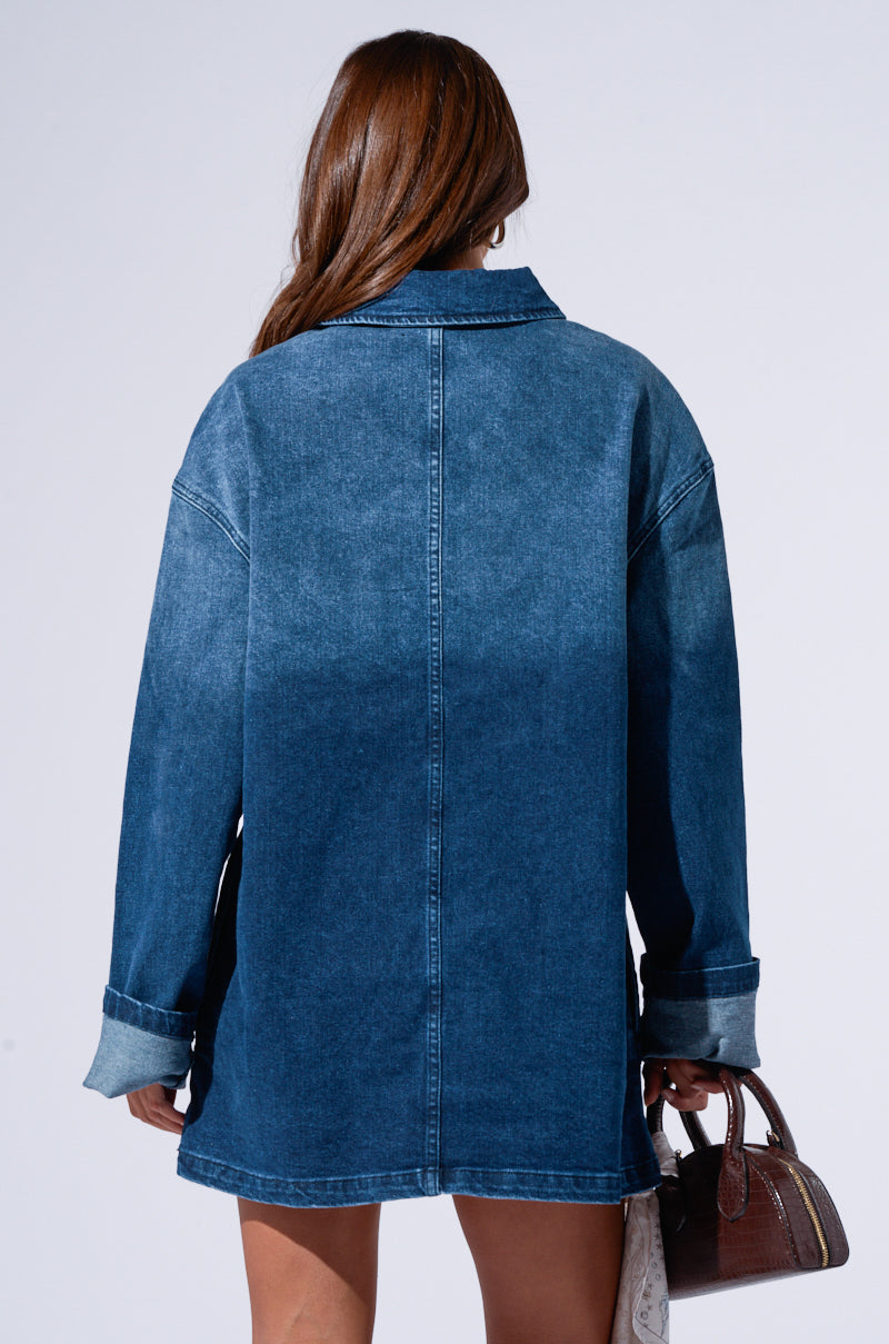 A person with long brown hair is facing away from the camera, wearing a COOL GIRL OVERSIZED DENIM SHIRT DRESS that exudes a casual-cute vibe. They are holding a small brown handbag in their right hand, set against a plain light gray background.
