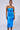 A woman stands against a plain background wearing the SHINE BRIGHT METALLIC BLUE MIDI DRESS, showcasing its shiny, metallic blue finish with a strapless bodycon silhouette and a midriff cut-out. Her hair is pulled back, and she looks confidently at the camera.