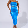 A woman stands confidently in the SHINE BRIGHT METALLIC BLUE MIDI DRESS, showcasing its strapless bodycon silhouette and small midriff cut-out under the bust. She pairs it with pointed-toe high heels featuring a checkered pattern. With her hair pulled back neatly, she strikes a pose with one hand on her hip against a plain background.