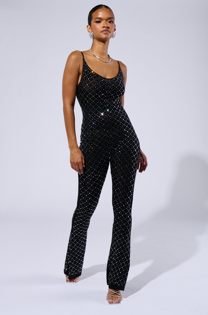 A person stands confidently in a black, form-fitting SABRINA RHINESTONE MESH JUMPSUIT adorned with small, shiny embellishments. They wear open-toed heels and minimal jewelry. The background is plain and well-lit, highlighting their party-perfect style.