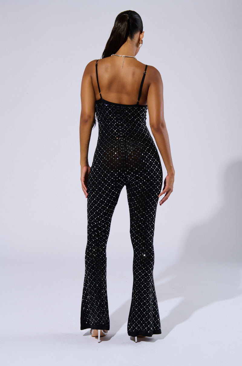 A woman with long hair in a ponytail is standing with her back to the camera, showcasing the form-fitting SABRINA RHINESTONE MESH JUMPSUIT. The sleeveless, black jumpsuit is adorned with small, sparkling studs and features a super stretchy material that flares at the bottom of the legs. She completes her look with high heels.