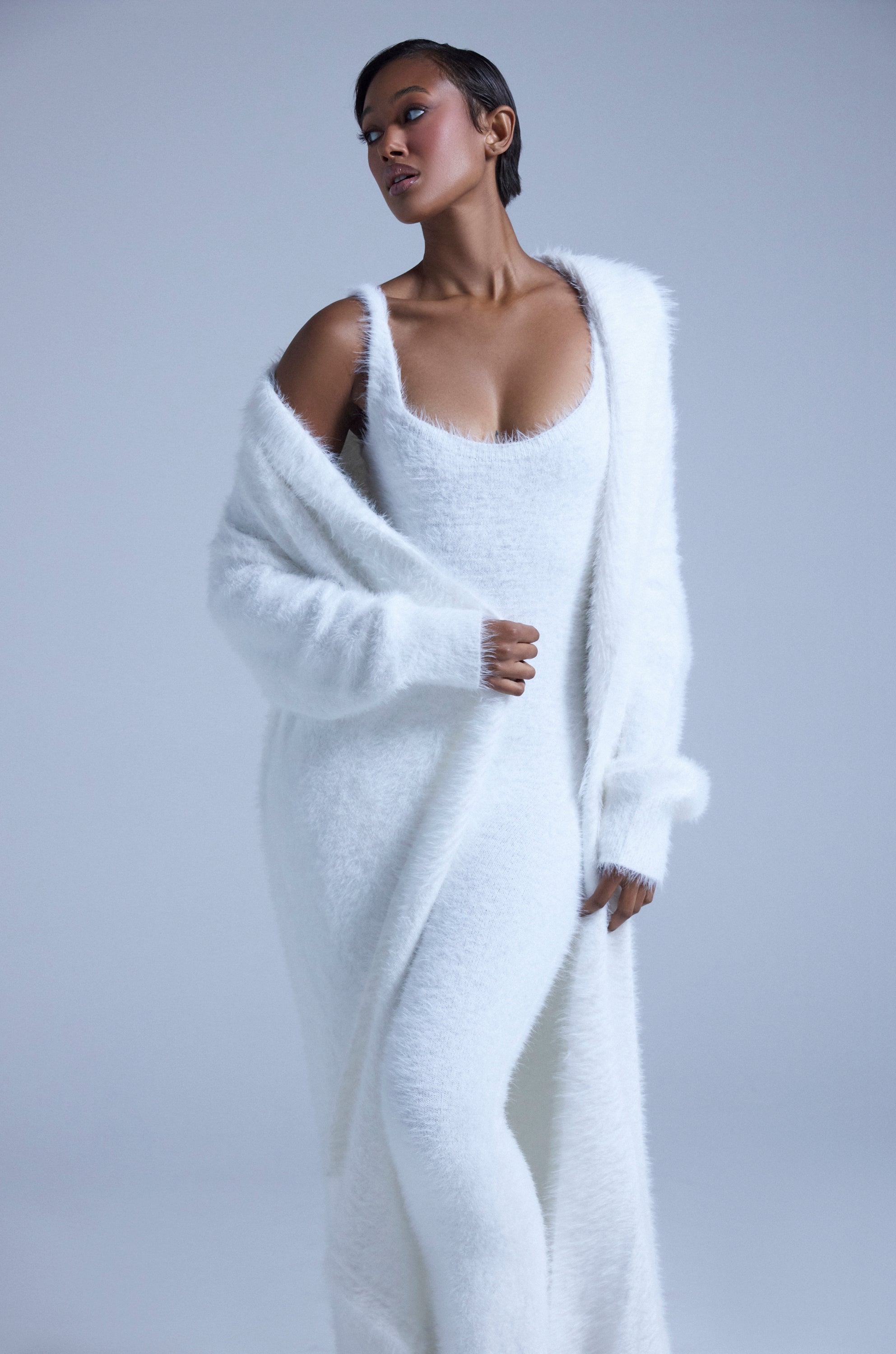 A woman models the FUZZY KNIT SHAWL NECK CARDIGAN over a long, fuzzy white dress, standing against a plain light grey background, gazing to the side with one arm slightly raised and the other by her side.