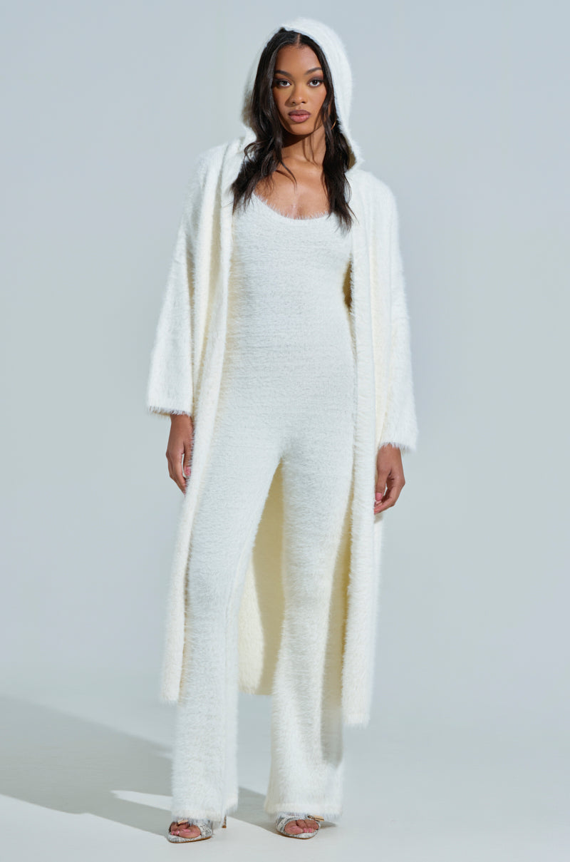 A woman stands confidently in a STAYING IN FUZZY KNIT JUMPSUIT, paired with a long hooded coat. Her long hair flows as she wears open-toed sandals, and the minimalistic background enhances her stylish loungewear look.