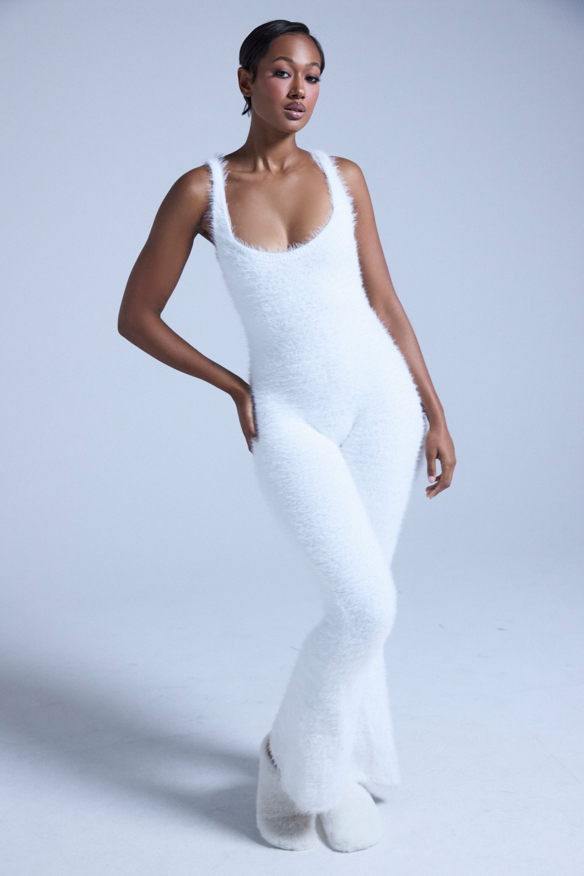 A woman stands confidently against a light gray background in the STAYING IN FUZZY KNIT JUMPSUIT, a fitted, cozy loungewear piece with a deep neckline—perfect for relaxing in style. She completes the look with matching fuzzy slippers. 0