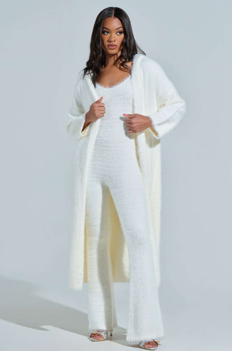 A person stands wearing the STAYING IN FUZZY KNIT JUMPSUIT in a fluffy white ensemble, featuring a long cardigan that epitomizes loungewear elegance. Open-toed heels add cozy sophistication against the plain light grey backdrop.