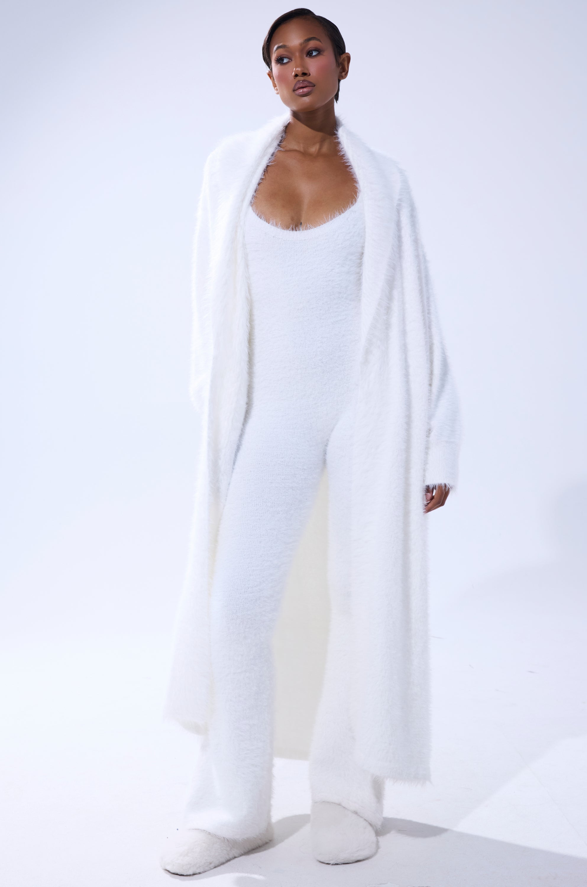 A woman stands against a white background wearing the STAYING IN FUZZY KNIT JUMPSUIT with a matching cardigan and white slippers. Her hair is pulled back, highlighting the cozy, soft, and textured look of her loungewear outfit. 2