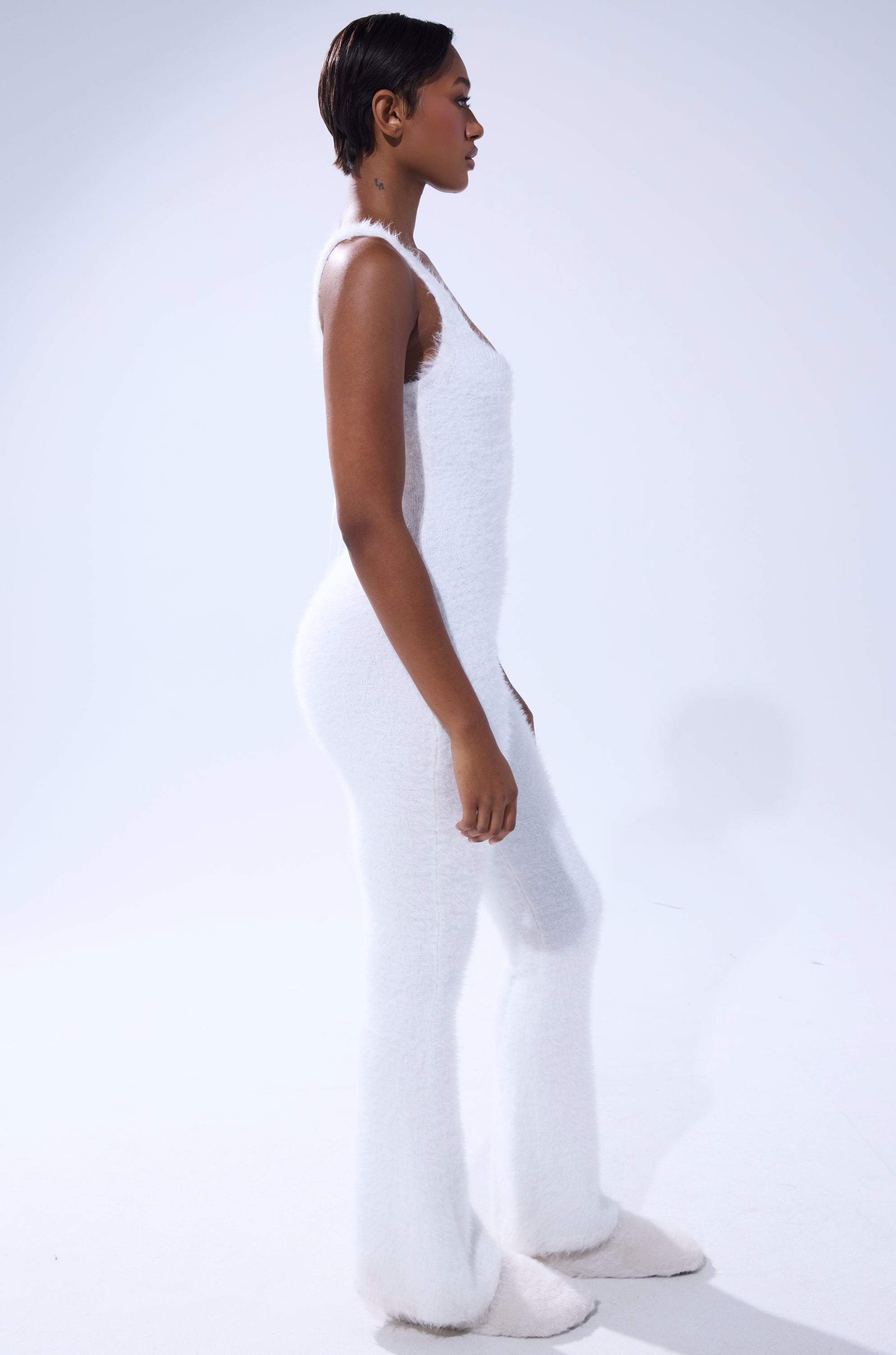 A person with short, close-cropped hair stands in profile against a white background, modeling the STAYING IN FUZZY KNIT JUMPSUIT—a fitted, sleeveless loungewear piece with flared legs and a soft, textured feel. 3