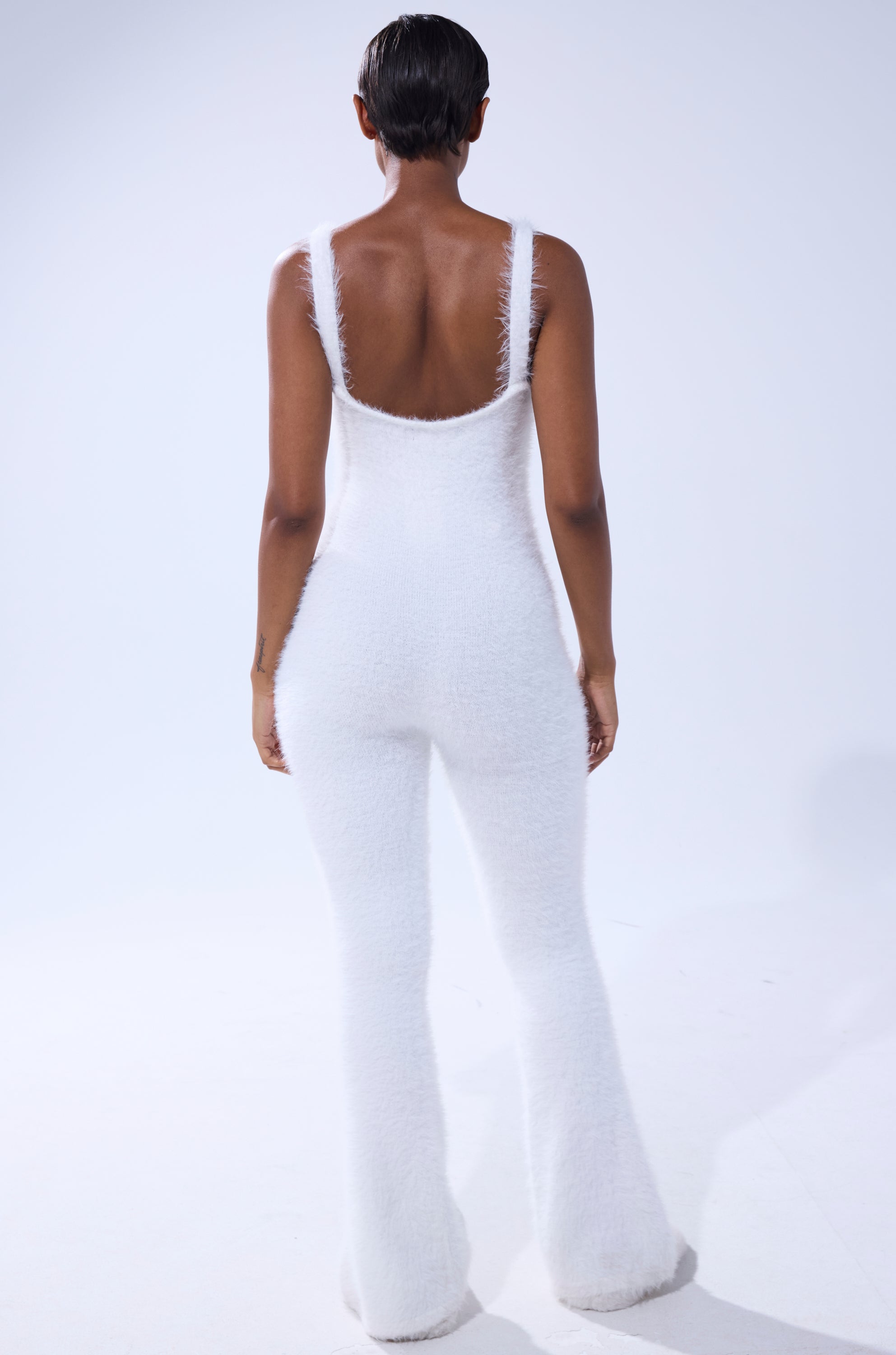 Facing away against a plain light background, a short-haired person models the STAYING IN FUZZY KNIT JUMPSUIT, featuring flared legs and a low back—an ideal choice for chic, comfortable loungewear. 4