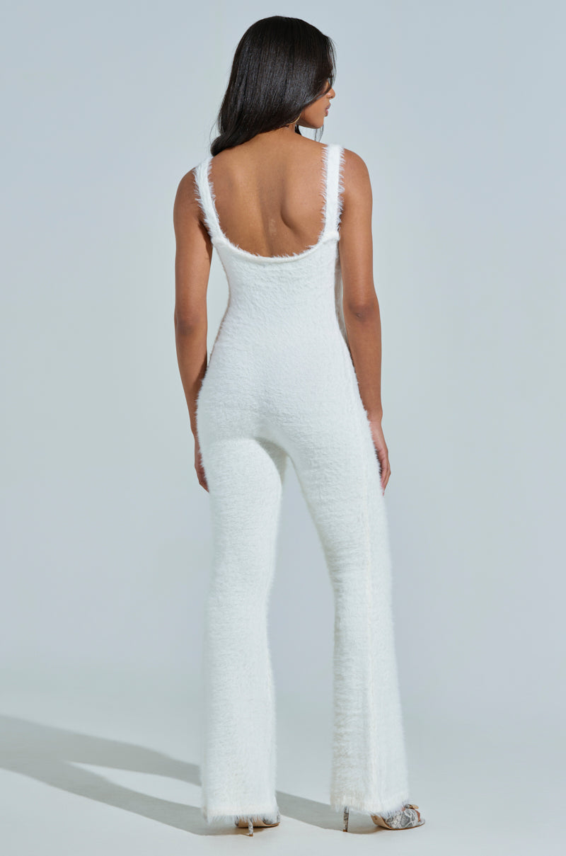 A woman with long dark hair models the STAYING IN FUZZY KNIT JUMPSUIT, a white, form-fitting loungewear piece featuring a low back and flared legs. She stands on a light-colored floor against a plain, light background, exuding effortless style and comfort.