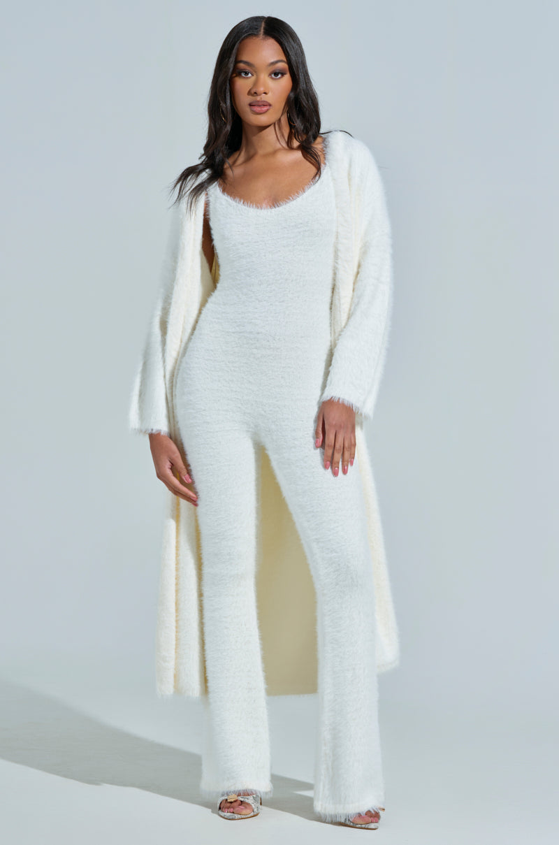 The woman dons the STAYING IN FUZZY KNIT JUMPSUIT in white with a matching long cardigan against a plain background. Her sleek, dark hair flows elegantly as she looks forward confidently, complementing her loungewear ensemble of flared pants and chic clear high-heeled shoes.