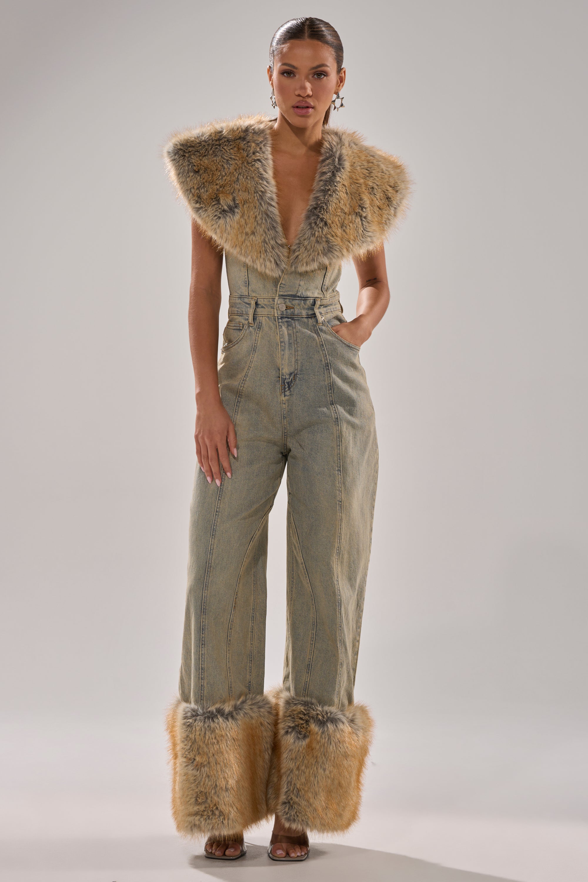 A woman wears the TALK THAT TALK DENIM FUR JUMPSUIT IN VINTAGE BLUE, a sleeveless, wide-leg jumpsuit with bold faux fur trim on the collar and cuffs, a deep V-neck, and high waist. She pairs it with heeled sandals against a plain light gray background.