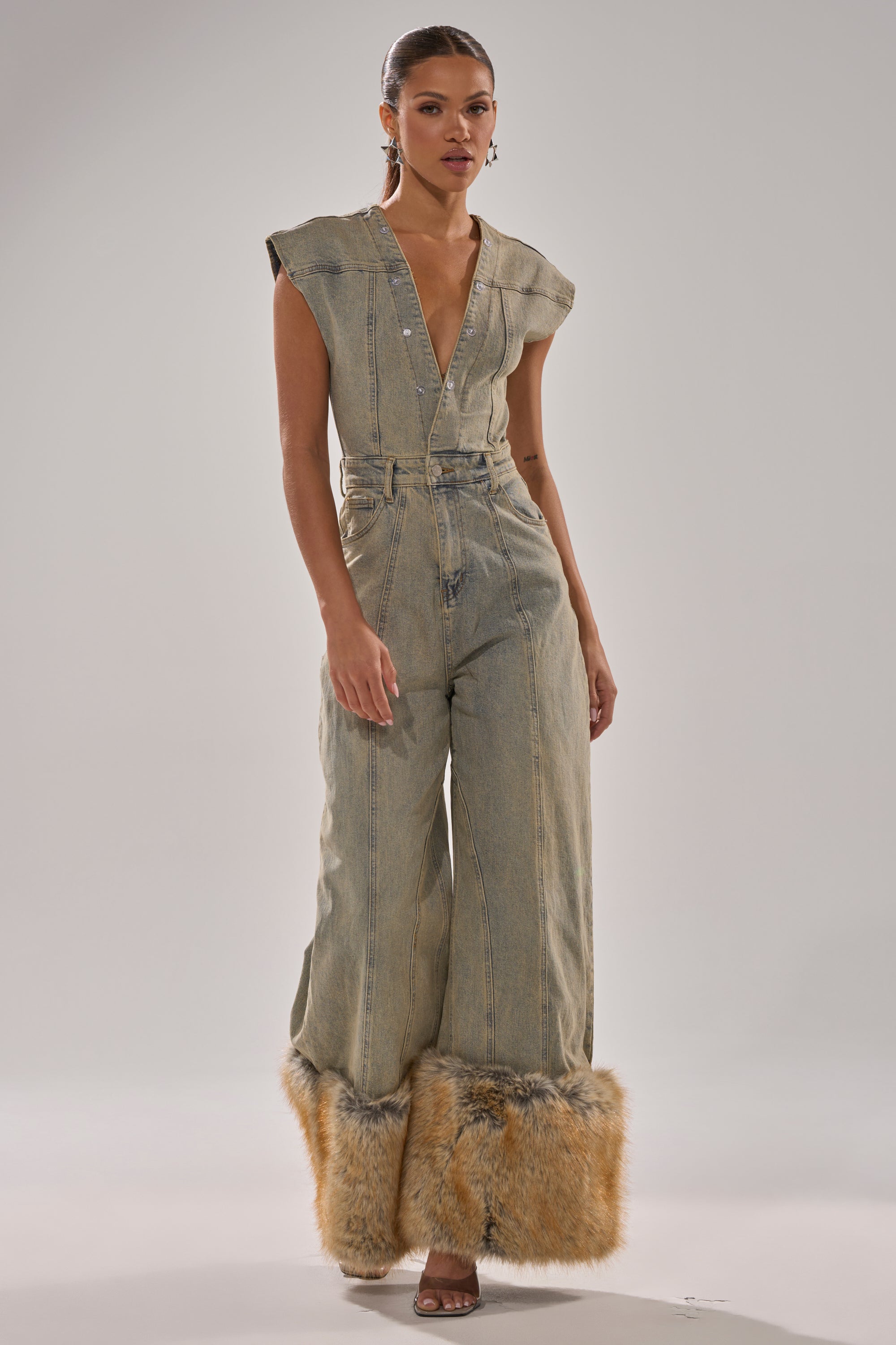 A woman stands in a studio wearing the TALK THAT TALK DENIM FUR JUMPSUIT IN VINTAGE BLUE, a deep V-neck, wide-leg denim piece trimmed with tan faux fur at the hems. Her hair is tied back and she pairs it with high-heeled sandals.