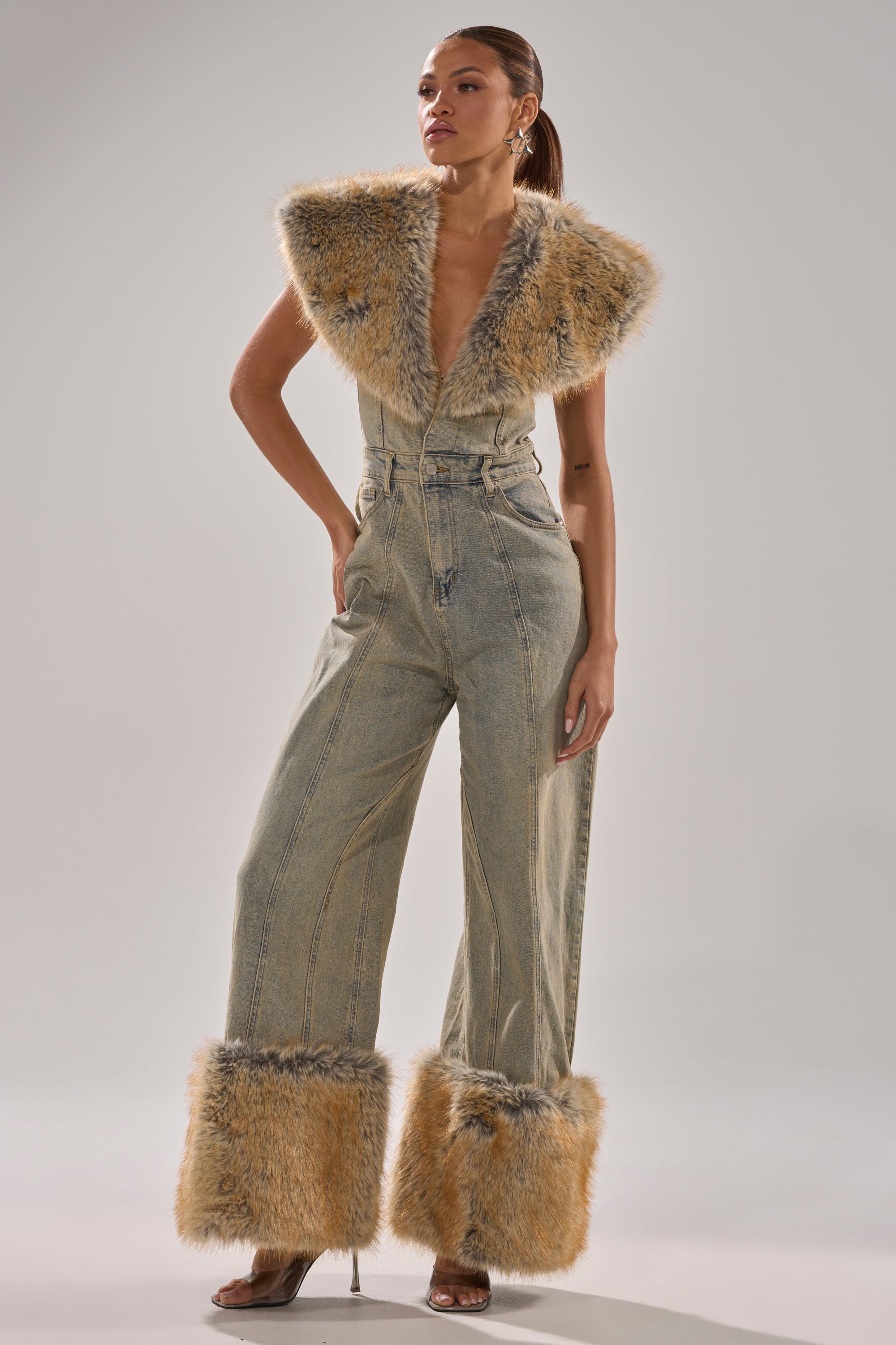 A woman models the TALK THAT TALK DENIM FUR JUMPSUIT IN VINTAGE BLUE, a wide leg denim jumpsuit with striking tan faux fur collar and ankle accents, blending rugged style with bold, soft details for a standout look.