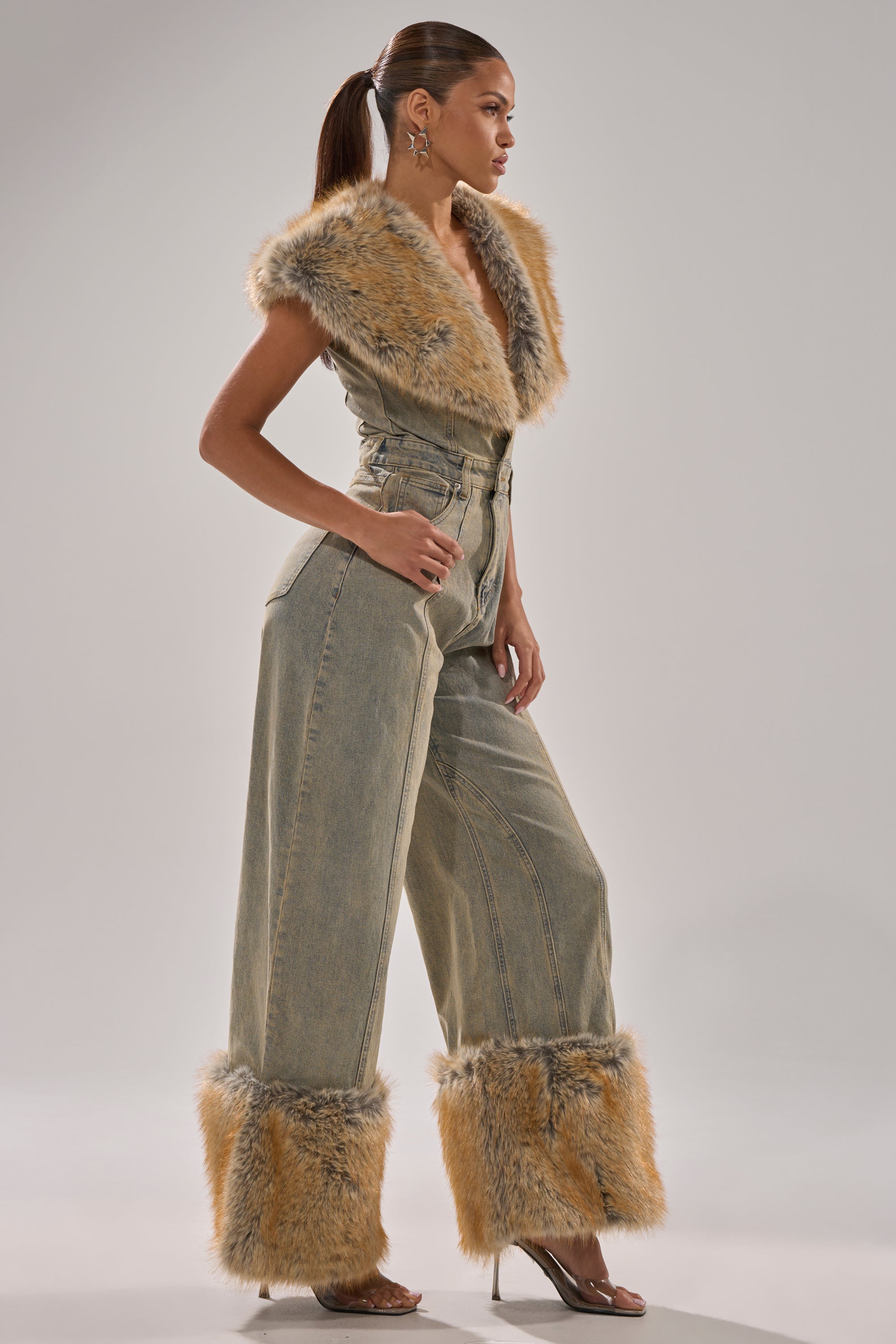 A woman in profile wears the TALK THAT TALK DENIM FUR JUMPSUIT IN VINTAGE BLUE, featuring wide legs and tan faux fur at the collar and cuffs. Her hair is in a ponytail and she pairs it with clear high-heeled shoes.