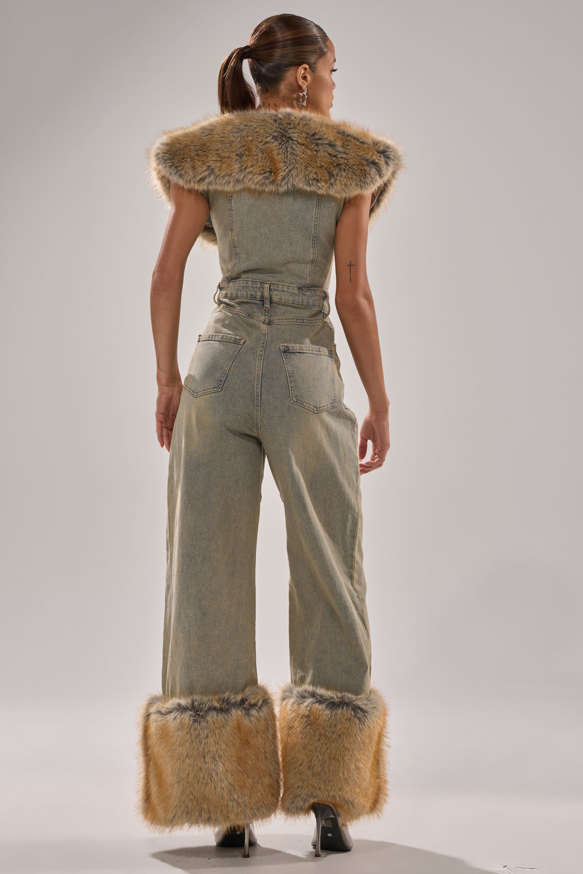 A woman stands facing away in the TALK THAT TALK DENIM FUR JUMPSUIT IN VINTAGE BLUE, featuring wide legs and fur trim on the collar and cuffs. Her hair is tied back as she poses against a plain light background.