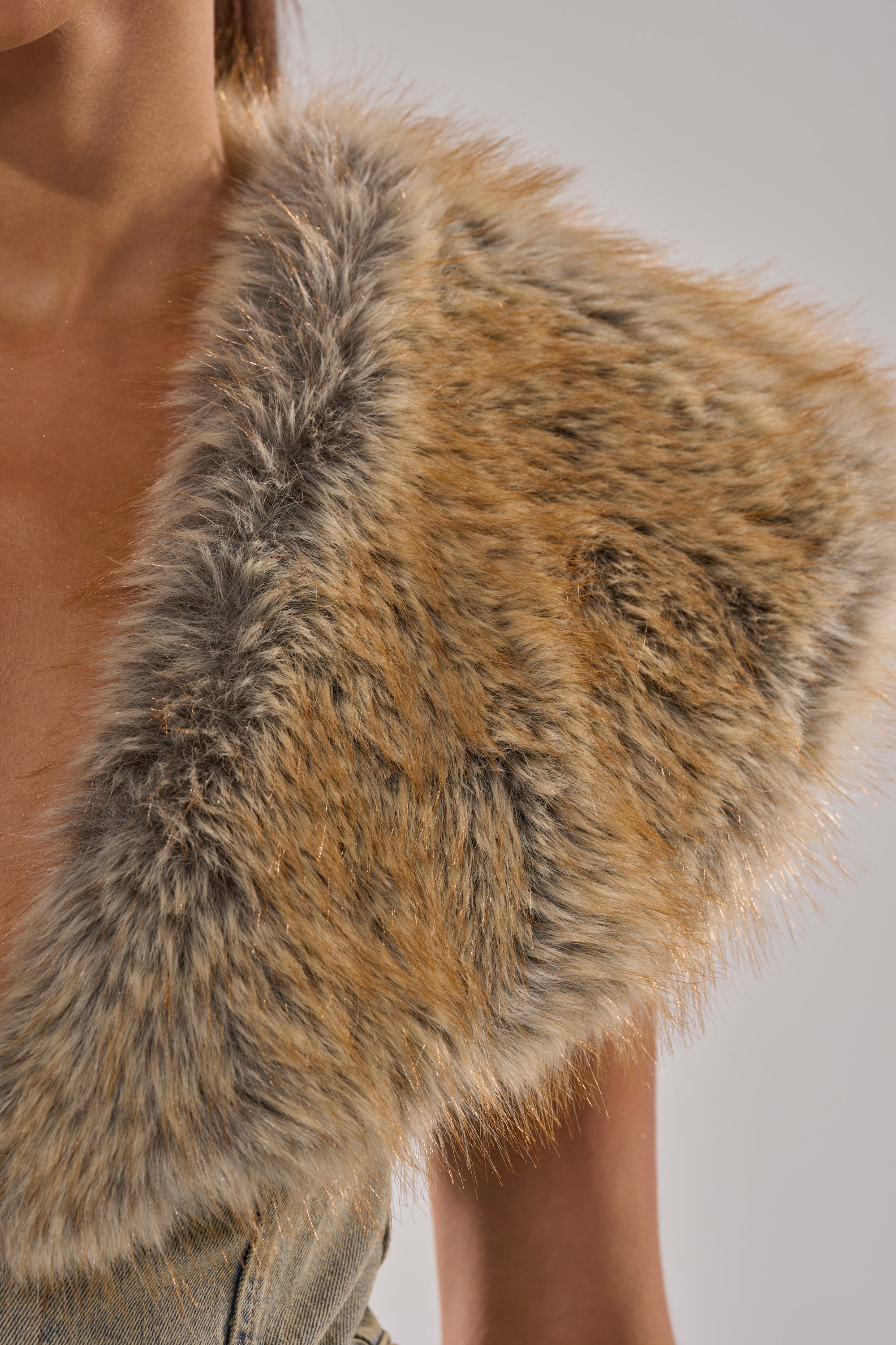 Close-up of a person wearing the TALK THAT TALK DENIM FUR JUMPSUIT IN VINTAGE BLUE, featuring a faux fur stole in brown, tan, and gray draped over one shoulder against a plain light background. Only the upper chest and shoulder are visible.