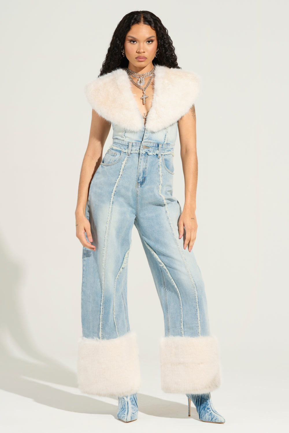 A person with long curly hair wears the chic TALK THAT TALK DENIM FUR JUMPSUIT, featuring a plunging V-neckline. This stylish outfit is completed with light blue high-waisted jeans embellished with white fur at the cuffs and patterned high-heeled boots, perfectly blending elegance and flair.