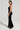 A woman in the HEY GIRL HEY VELVET JUMPSUIT poses against a plain background, showcasing its sleek and stretchy fabric. The jumpsuit features a halter neck and an open back design. She accessorizes with hoop earrings and black high heels, standing confidently with one hand on her hip.