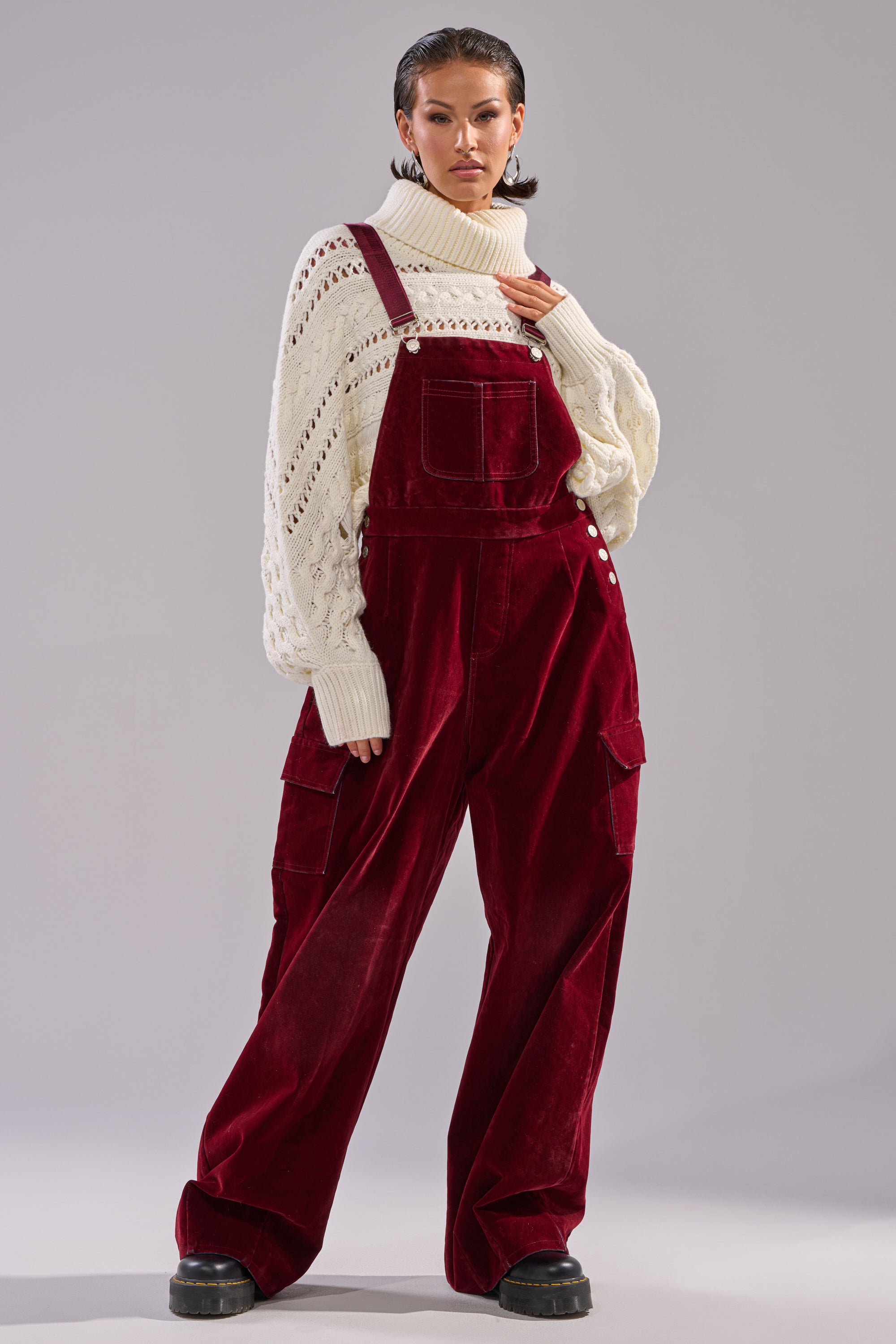 A person poses confidently in IT'S GIVING VINTAGE VELVET DENIM OVERALLS—a wide-leg silhouette with cargo pockets—layered over a chunky cream turtleneck and paired with black platform shoes, against a plain light gray backdrop.