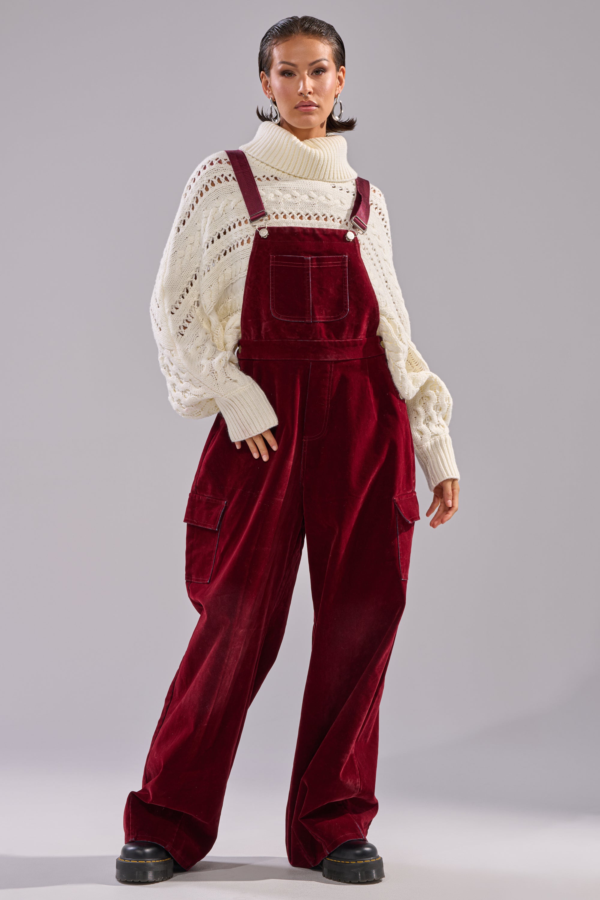 Someone wearing the IT'S GIVING VINTAGE VELVET DENIM OVERALLS with a wide-leg silhouette and a chunky white turtleneck stands confidently against a plain gray background.