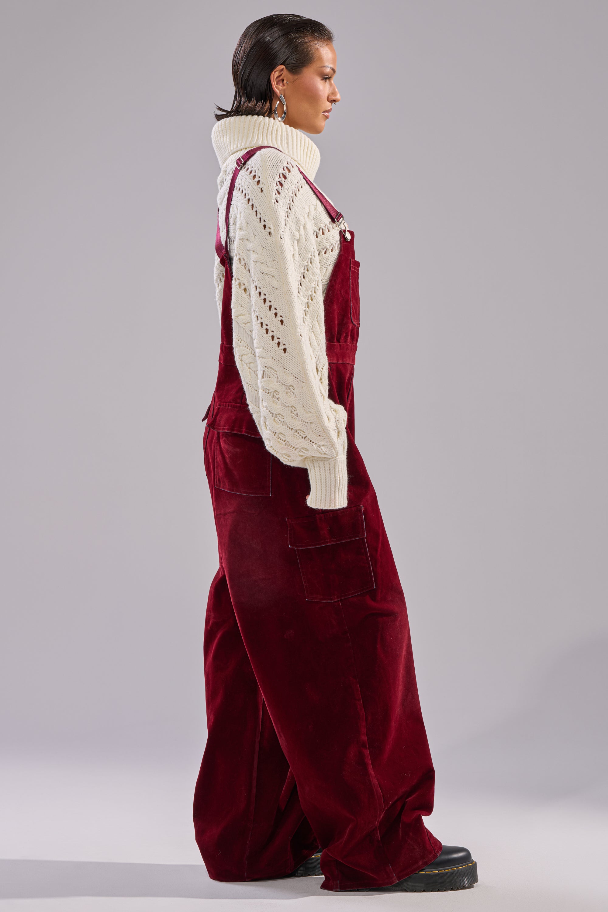 Someone stands sideways in a white knit turtleneck layered under IT'S GIVING VINTAGE VELVET DENIM OVERALLS with a wide-leg fit, short brown hair, and black platform shoes, set against a plain gray background.