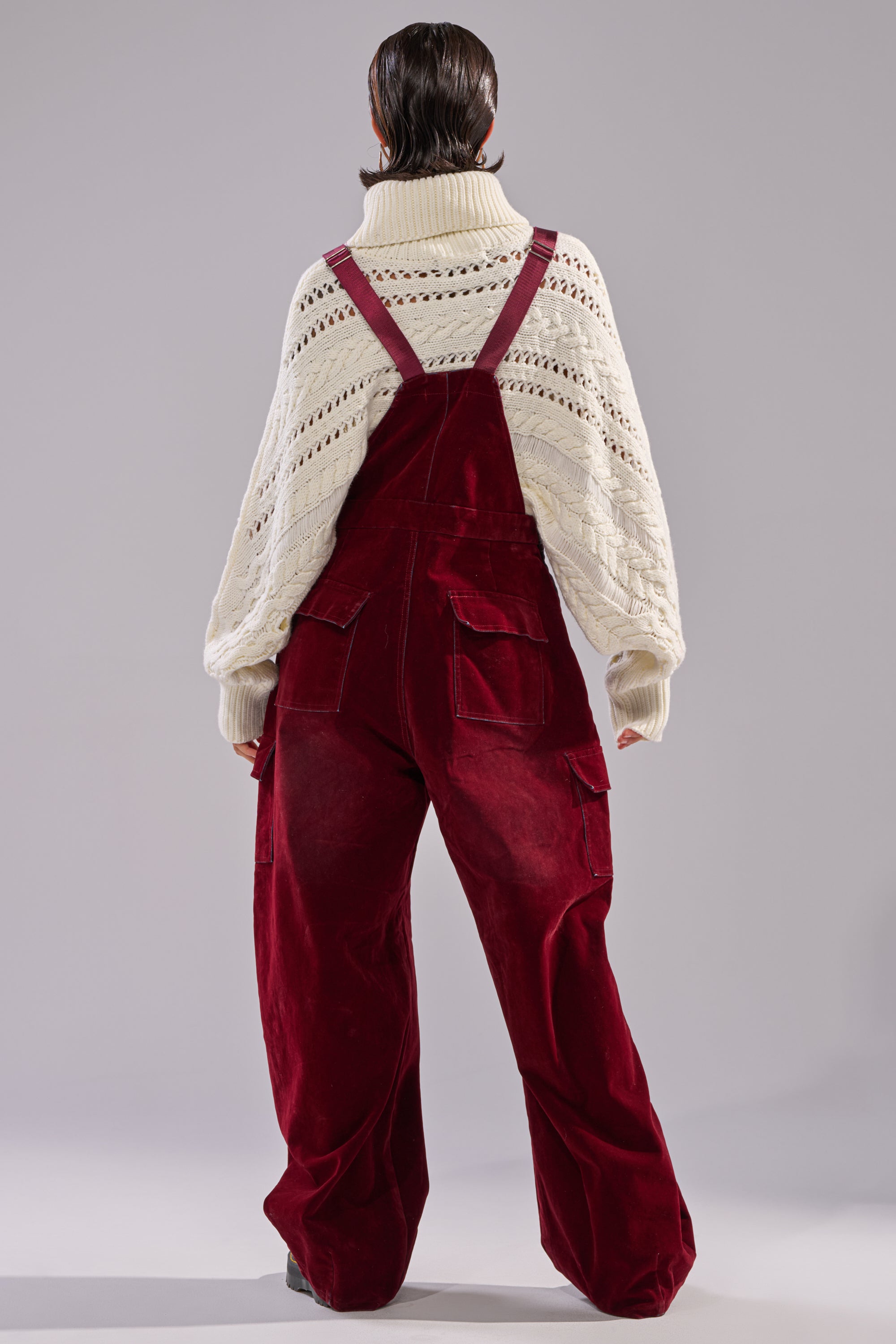 A person faces away, wearing a chunky white turtleneck and the IT'S GIVING VINTAGE VELVET DENIM OVERALLS with a wide-leg silhouette, set against a plain gray background.