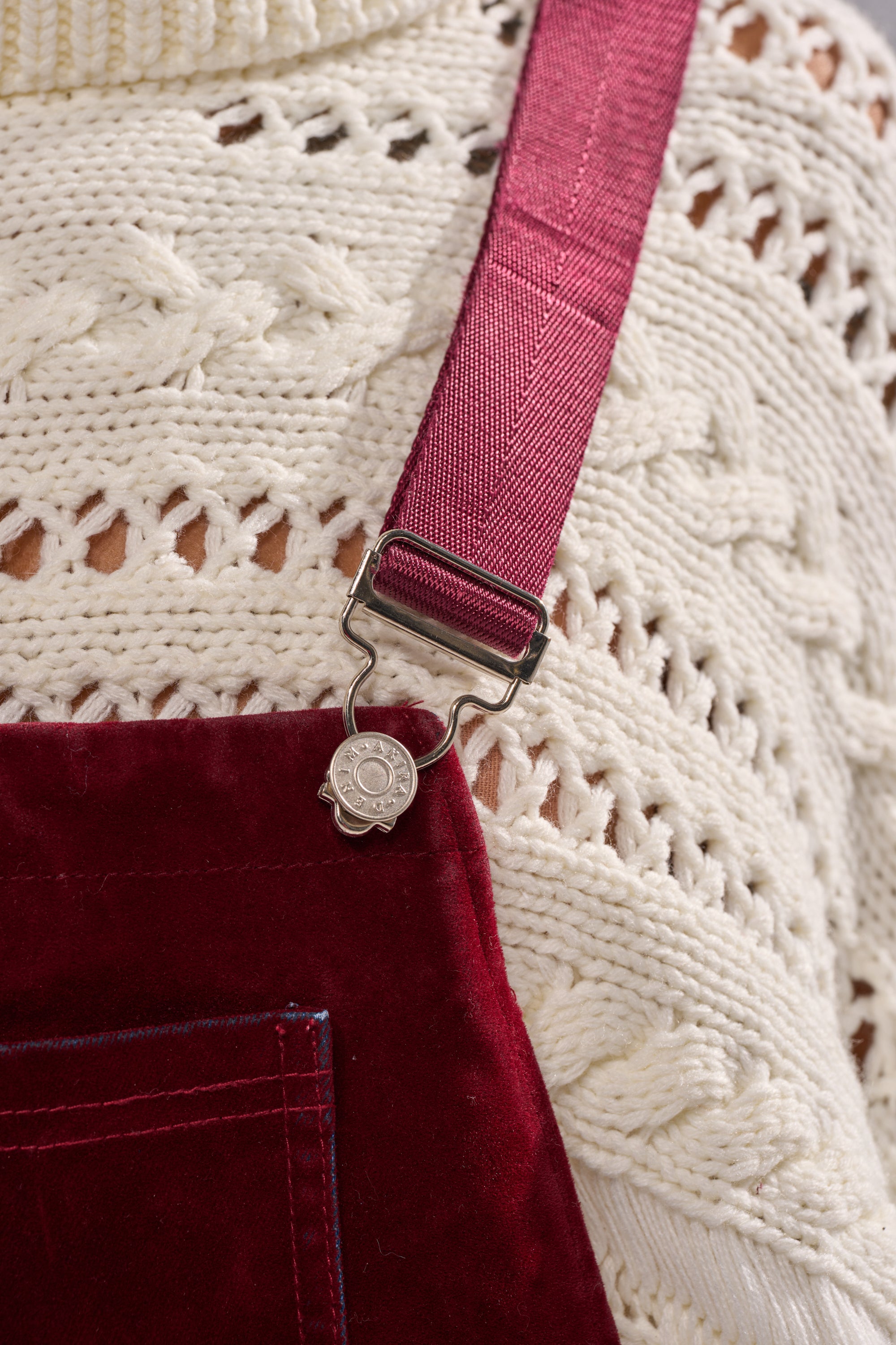 Close-up of a person wearing a white knit sweater and IT'S GIVING VINTAGE VELVET DENIM OVERALLS, featuring a red hue, silver clasp and button at the strap, and a chic wide-leg silhouette.