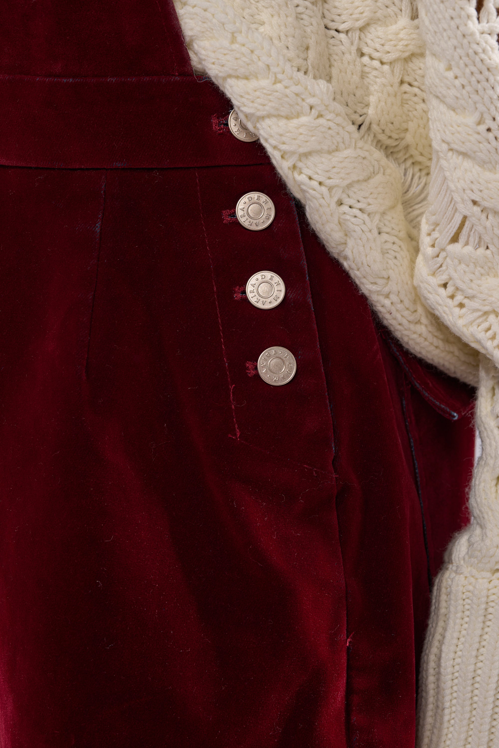 A person wears IT'S GIVING VINTAGE VELVET DENIM OVERALLS in deep red with silver buttons and utility pockets, paired with a thick cream cable-knit sweater draped at the waist.