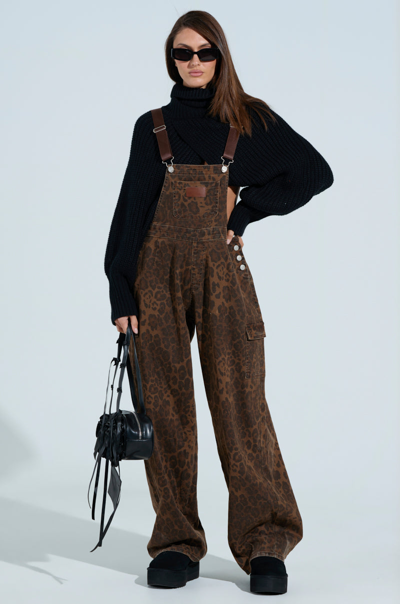 A person in PULL UP ON ME leopard print overalls with adjustable buckled straps, a black turtleneck sweater, and black platform shoes holds a small black handbag. With long hair and sunglasses, they stand against a light background.