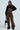 A person in PULL UP ON ME leopard print overalls with adjustable buckled straps, a black turtleneck sweater, and black platform shoes holds a small black handbag. With long hair and sunglasses, they stand against a light background.