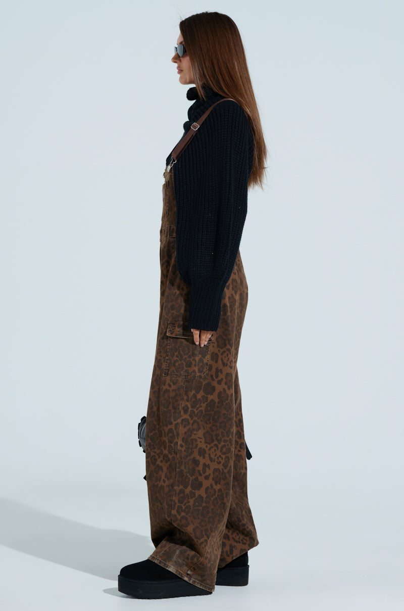A person with long brown hair stands in profile wearing PULL UP ON ME LEOPARD OVERALLS, featuring adjustable buckled straps and cargo pockets, paired with a black turtleneck sweater and platform shoes. Sunglasses complement the look as they face sideways against a plain background.
