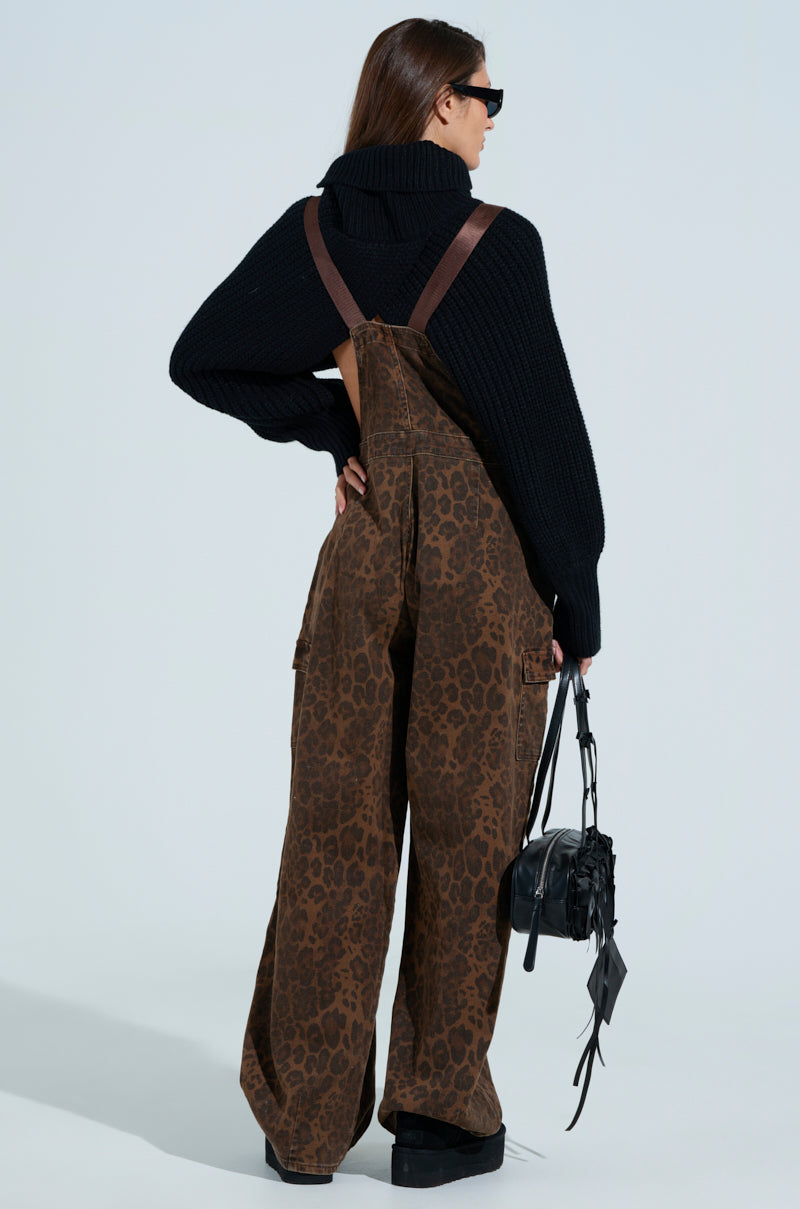A person with long hair wears a black turtleneck sweater and PULL UP ON ME LEOPARD OVERALLS, characterized by adjustable buckled straps. Black boots complement their look, while a tassel-adorned black bag hangs from their shoulder. They stand against a plain background, facing away from the camera.