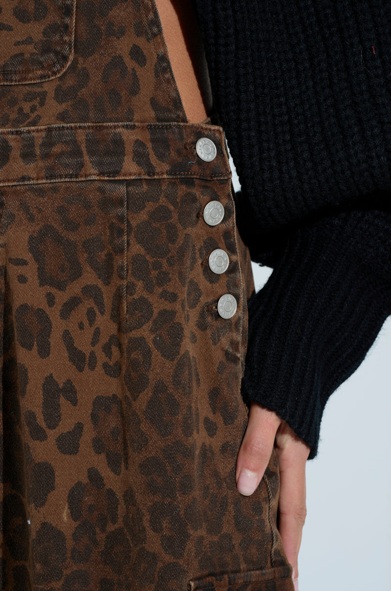 A close-up of someone in PULL UP ON ME LEOPARD OVERALLS with silver buttons and adjustable buckled straps, paired with a black knit sweater, hand resting on the side.