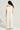 A person with long, curly hair is wearing the "YA YOU THOUGHT FRENCH TERRY JUMPSUIT," an off-the-shoulder piece in light gray made from a stretchy cotton blend. They stand facing away, showcasing the wide-leg silhouette of the jumpsuit against a plain, light background.