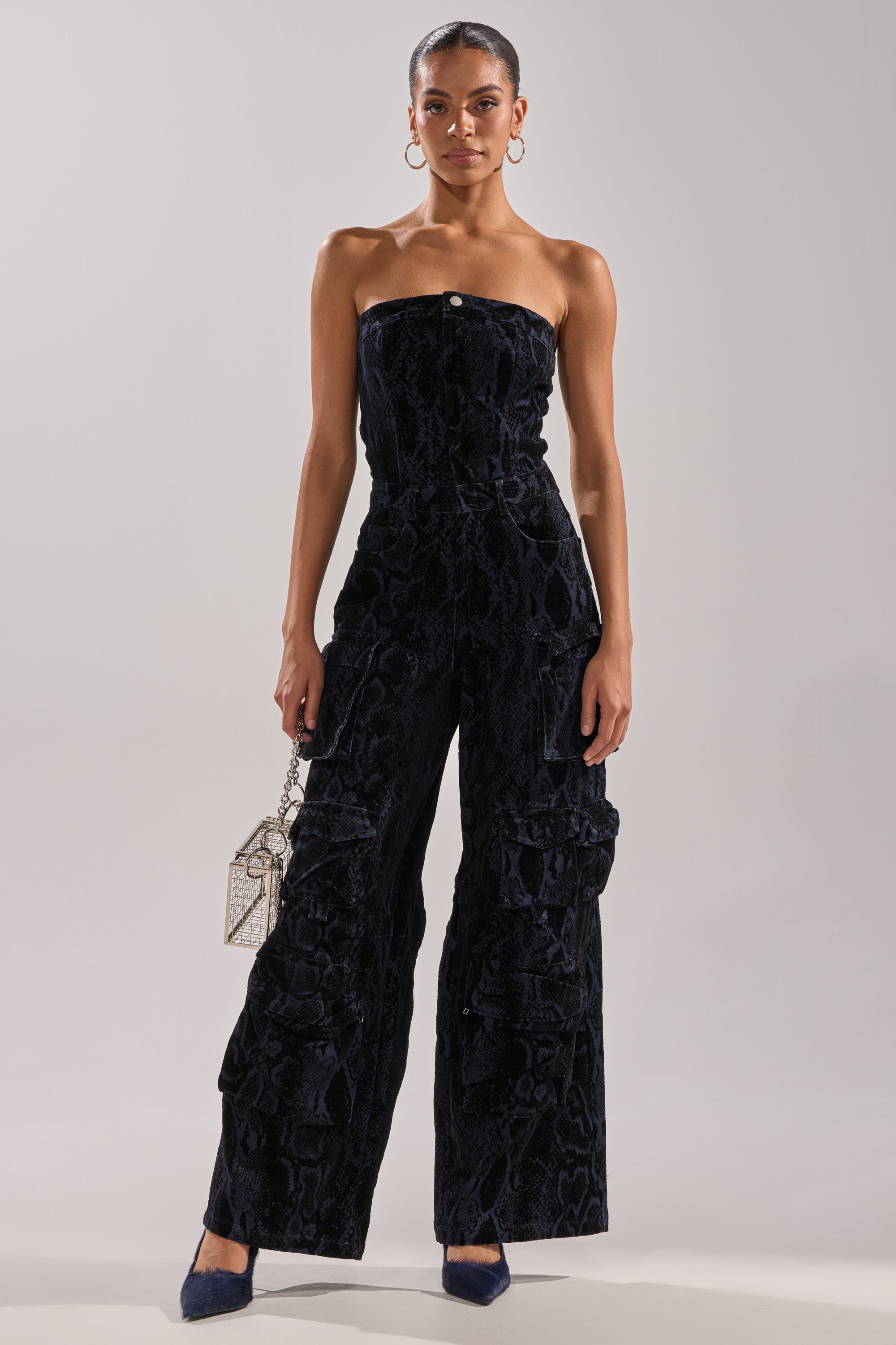 A woman faces forward in the CAUGHT YOUR EYE VELVET DENIM JUMPSUIT IN SNAKEPRINT with a textured black finish and wide legs. She accessorizes with a small silver handbag, hoop earrings, pulled-back hair, and pointed-toe heels.