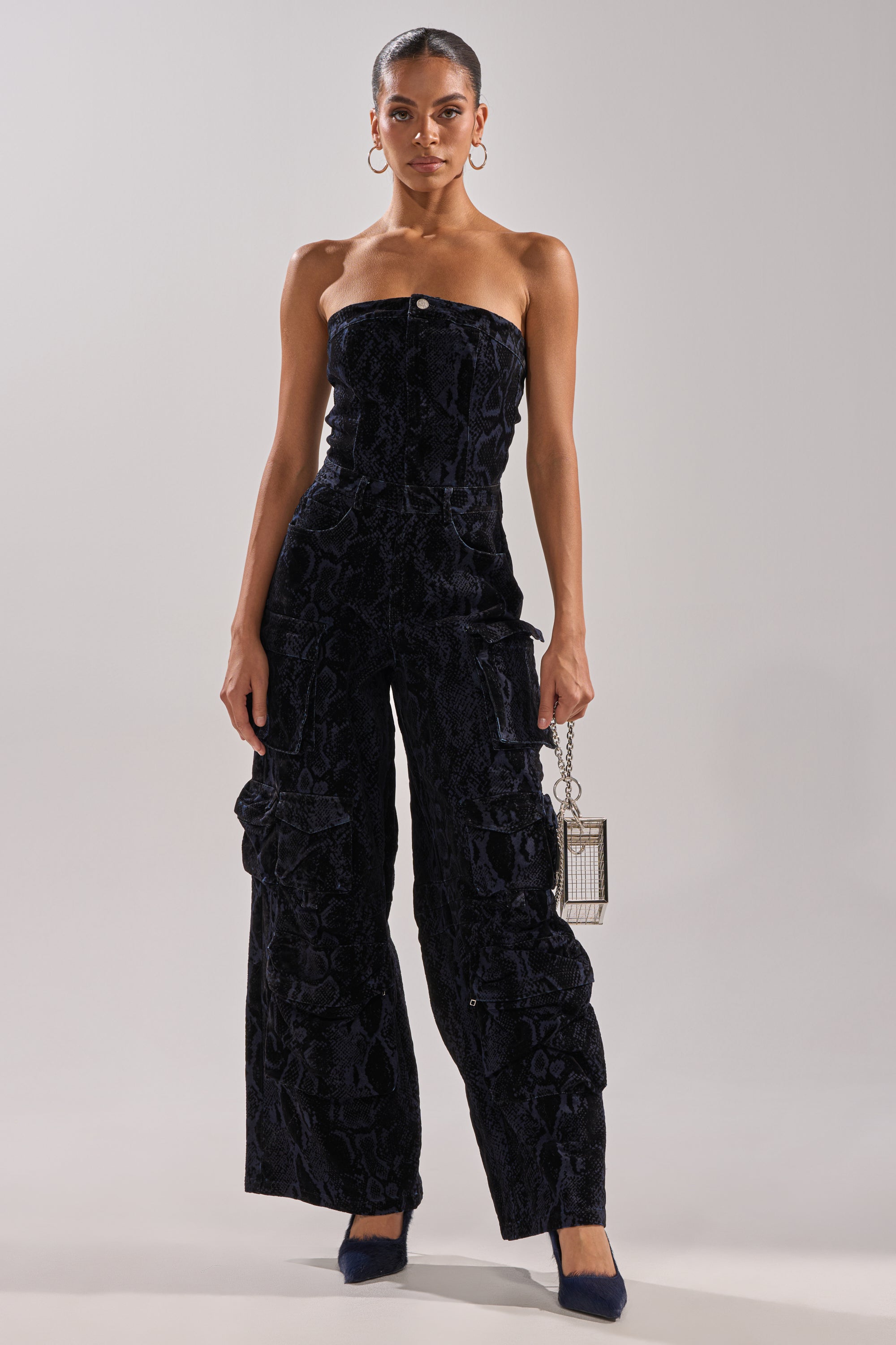 A woman poses against a plain backdrop in the CAUGHT YOUR EYE VELVET DENIM JUMPSUIT IN SNAKEPRINT, featuring wide legs. She accessorizes with a metallic handbag, hoop earrings, and dark heels, wearing her hair pulled back for a stylish touch.