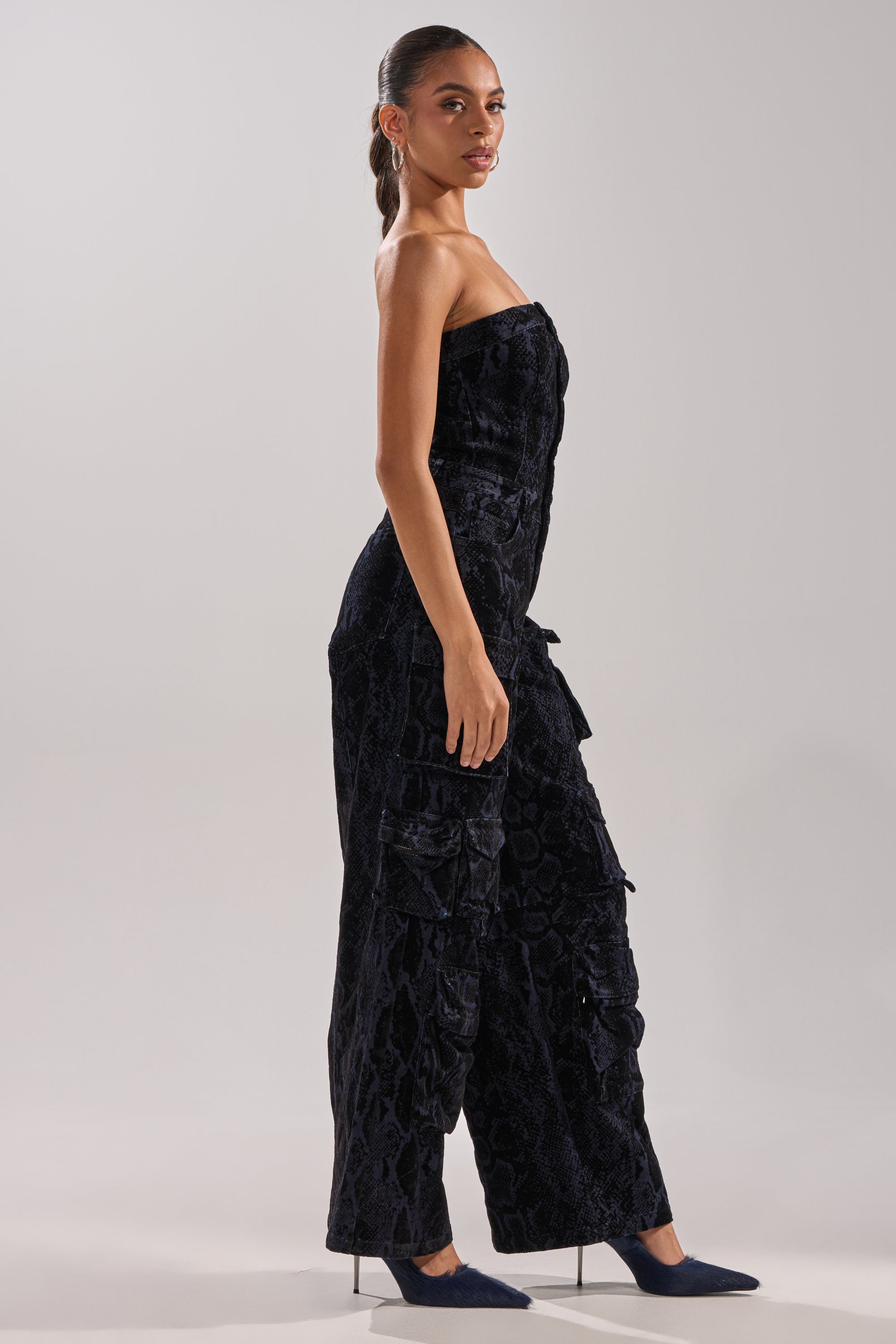 A woman stands sideways, facing the camera against a plain light background, wearing the CAUGHT YOUR EYE VELVET DENIM JUMPSUIT IN SNAKEPRINT and heeled shoes.