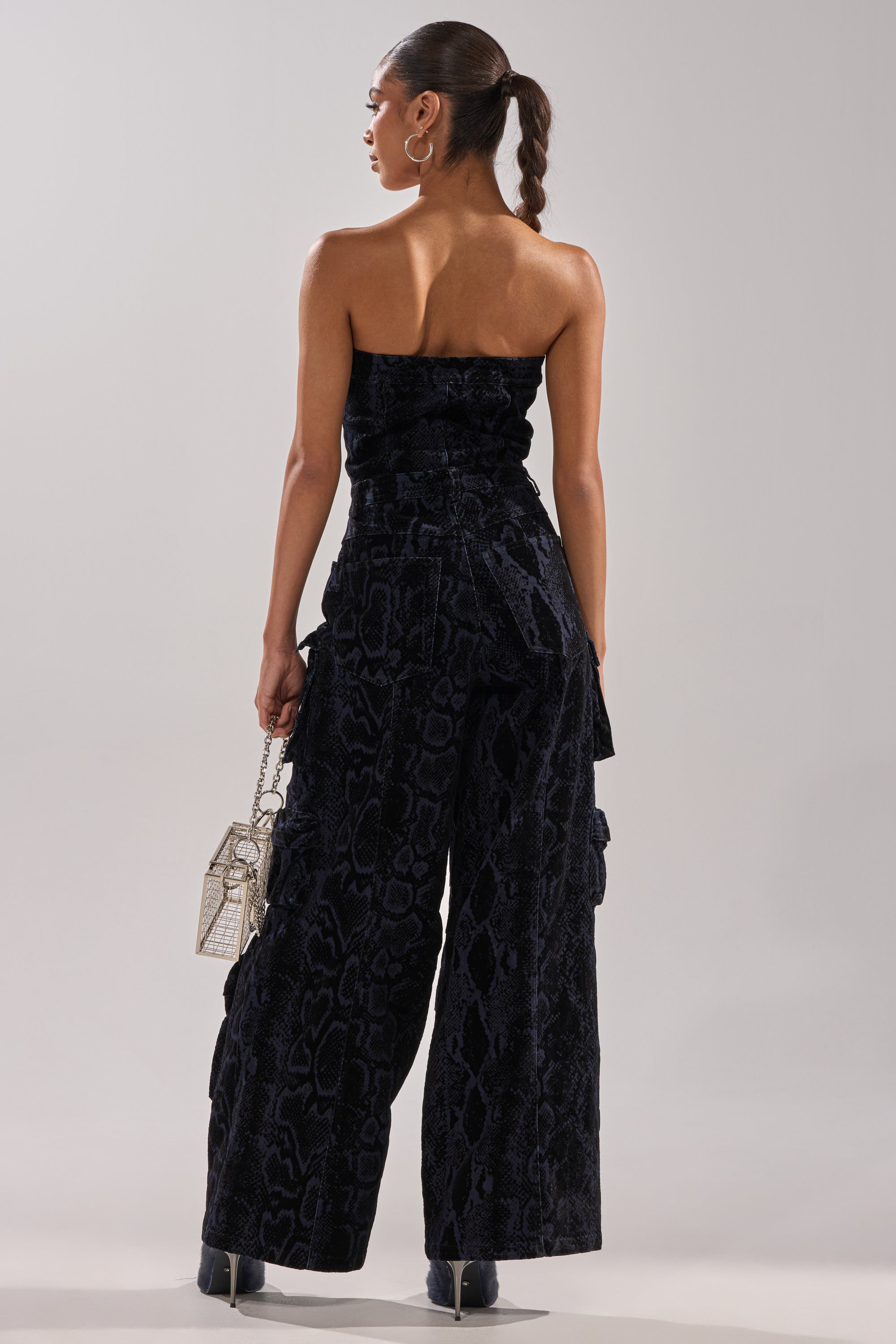 A woman stands facing away, wearing the CAUGHT YOUR EYE VELVET DENIM JUMPSUIT IN SNAKEPRINT with wide legs. She holds a metallic handbag, wears hoop earrings, and her hair is styled in a sleek ponytail.