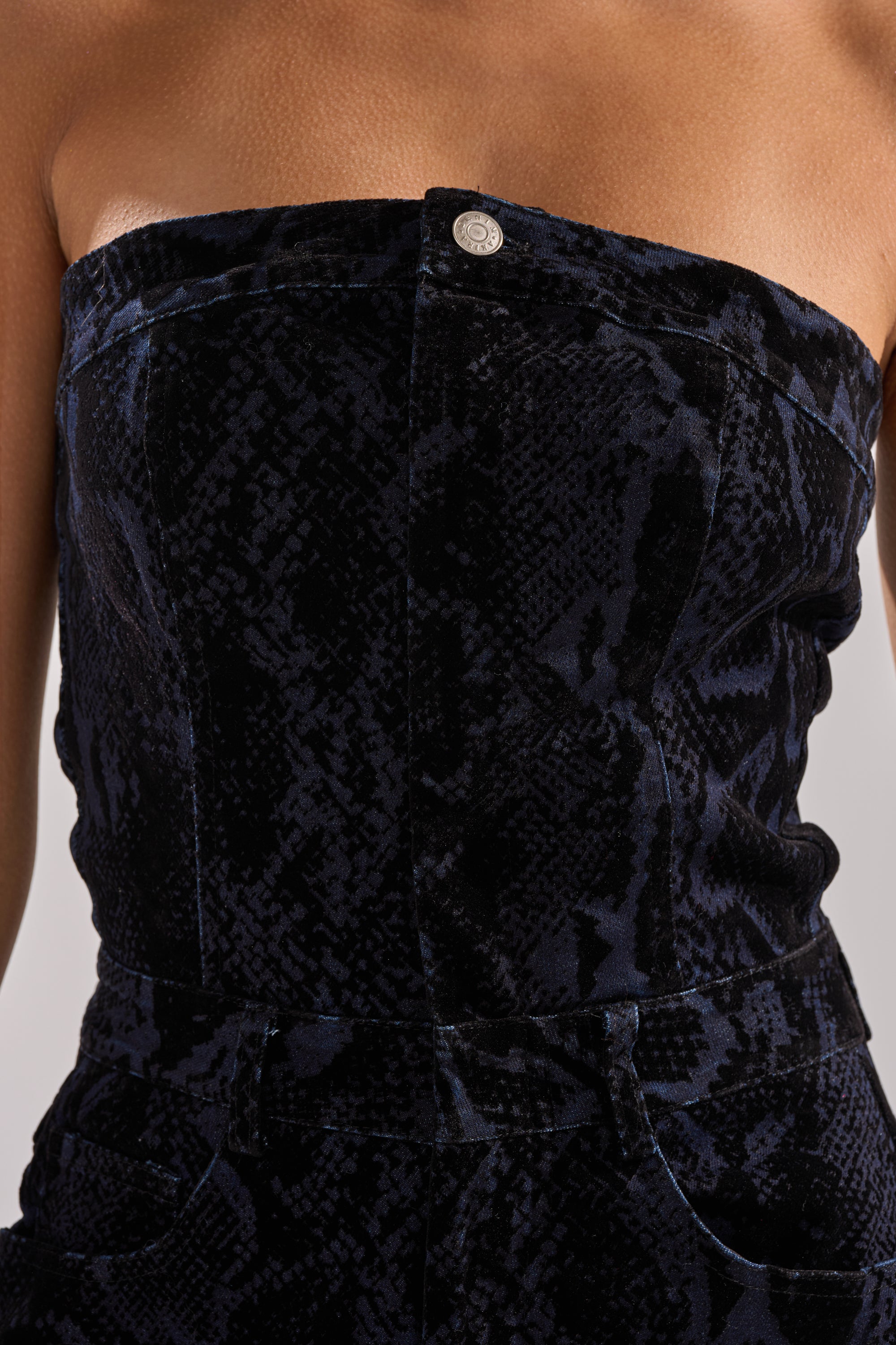 A close-up of a person wearing the CAUGHT YOUR EYE VELVET DENIM JUMPSUIT IN SNAKEPRINT, featuring a strapless design, dark velvet with subtle snake print, and a visible metal button at the top center.