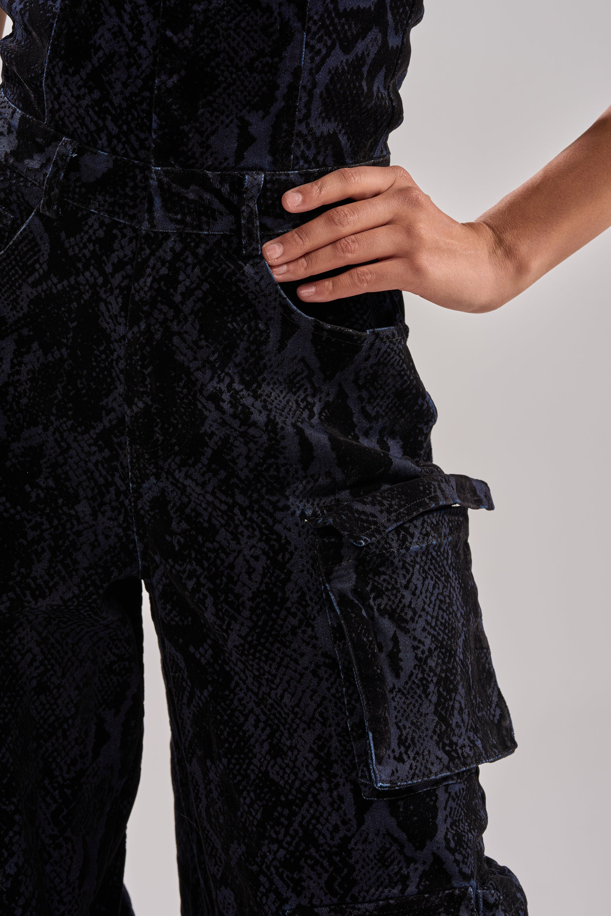 A person with one hand on their hip models the CAUGHT YOUR EYE VELVET DENIM JUMPSUIT IN SNAKEPRINT, featuring dark snakeprint jumpsuit pants with a cargo pocket, shown waist to mid-thigh against a plain background.