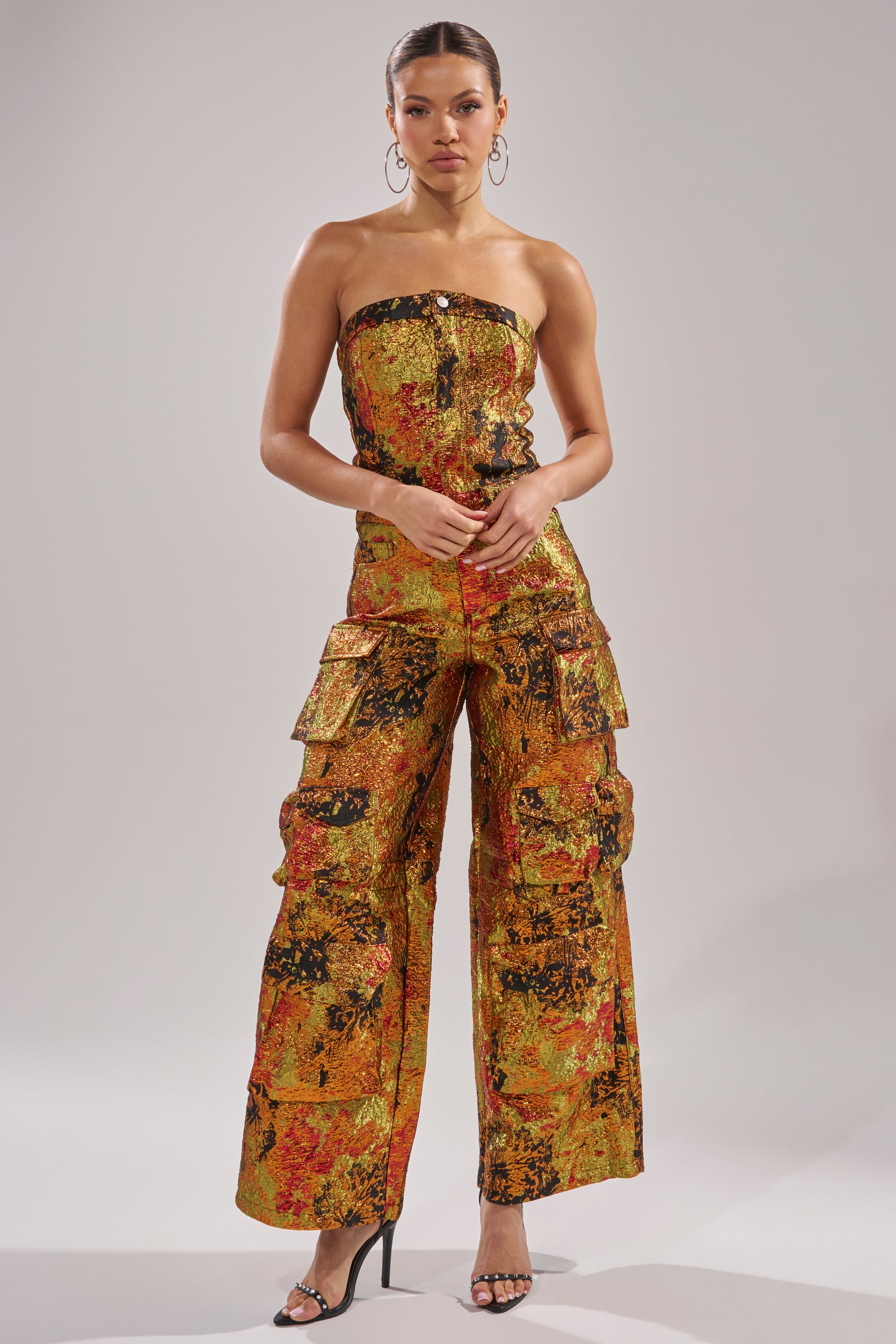 A woman poses against a plain backdrop in the NIKKI BROCADE JUMPSUIT, a strapless, wide-leg jumpsuit with cargo pockets and a bold orange, yellow, and black abstract print. She completes the look with hoop earrings and black heeled sandals.