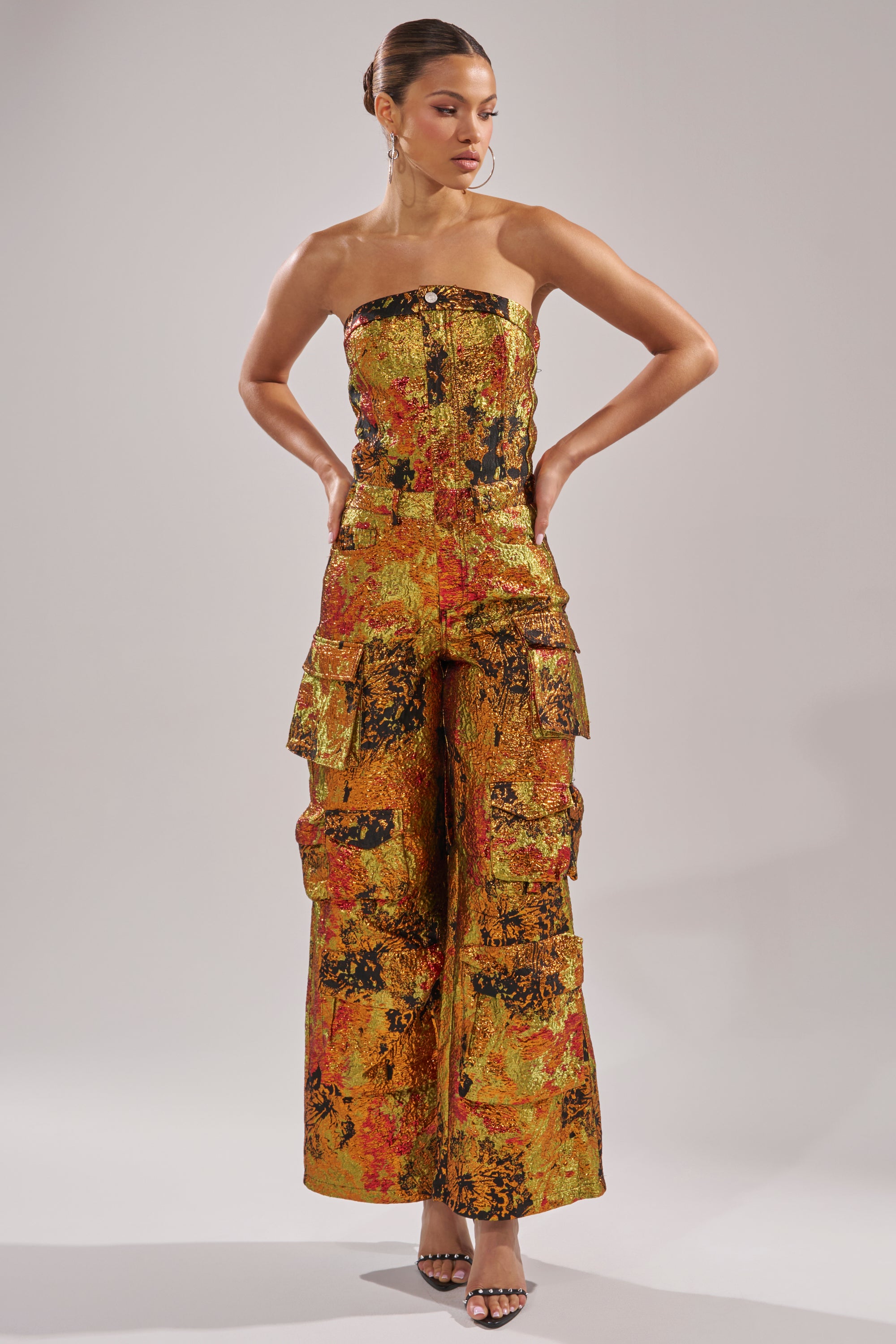 Wearing the NIKKI BROCADE JUMPSUIT, a woman stands with hands on hips, gazing to the side. The strapless metallic brocade jumpsuit features wide legs and multiple pockets. She completes the look with black heels and a sleek updo.