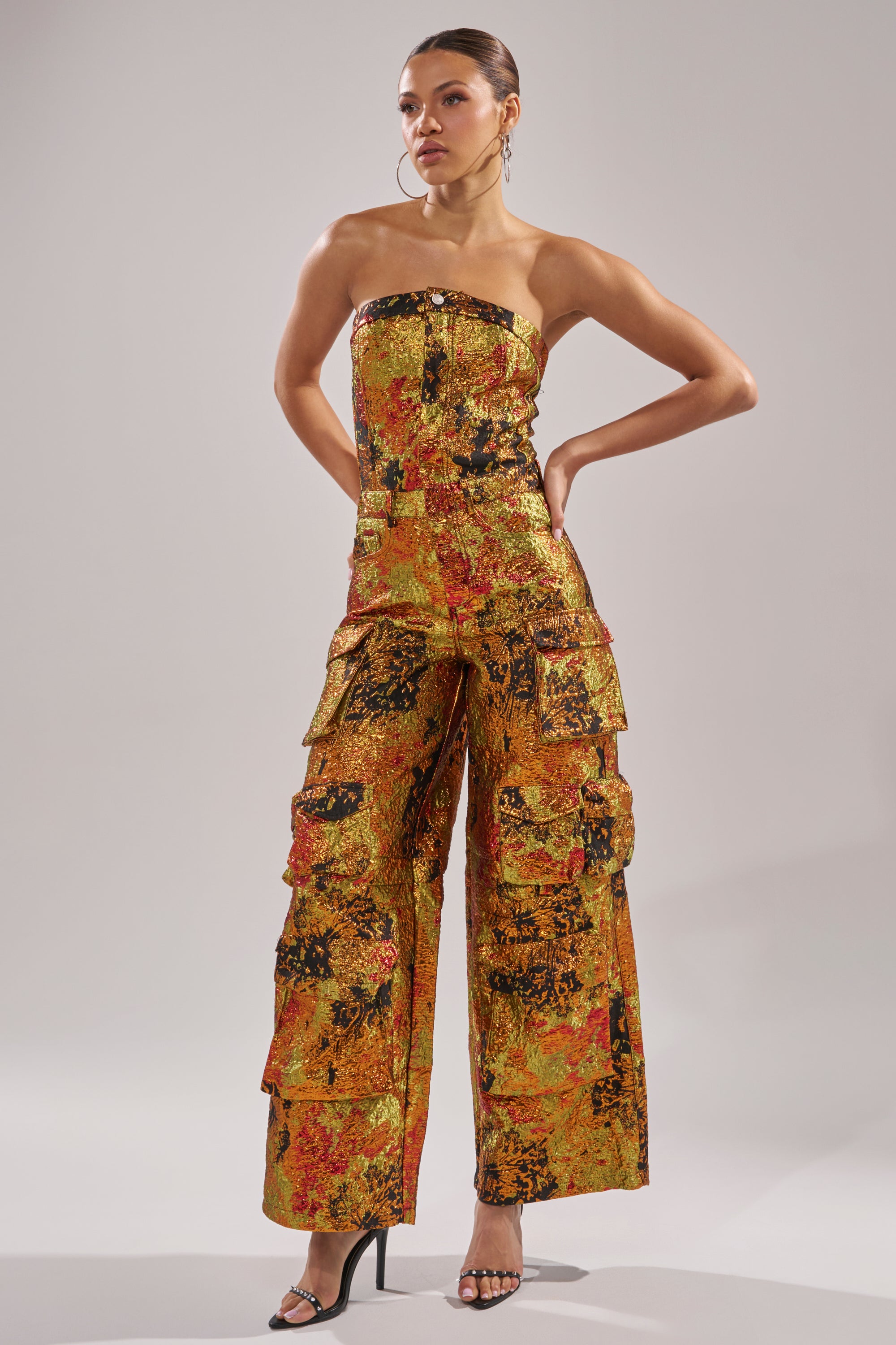 A woman stands confidently in the NIKKI BROCADE JUMPSUIT, a strapless eveningwear piece featuring a wide-leg silhouette, abstract orange, yellow, and black pattern, multiple pockets, and paired with black high-heeled sandals.