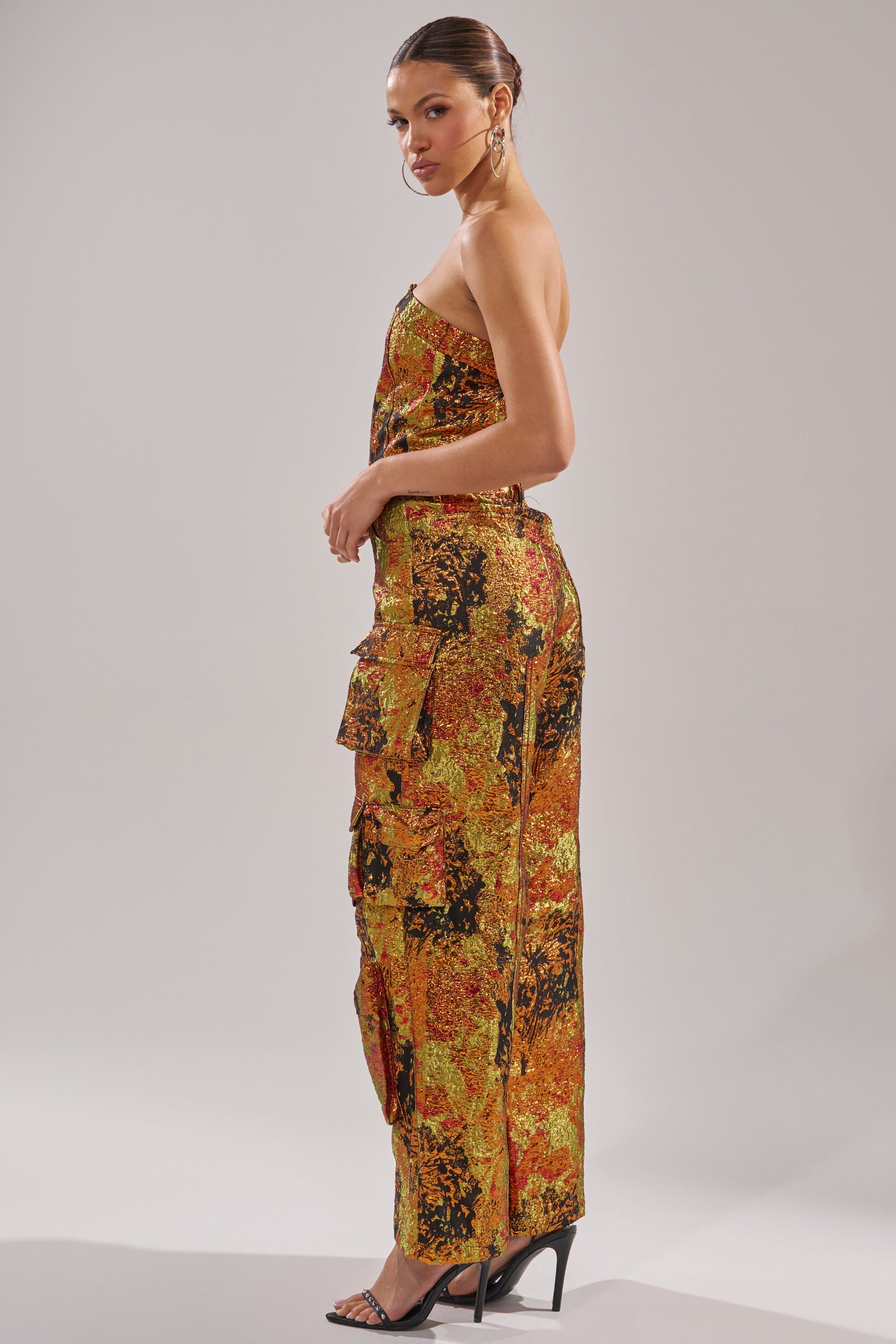 A woman stands sideways in the strapless NIKKI BROCADE JUMPSUIT with wide legs and side pockets, her hair in a sleek bun. She completes the look with hoop earrings and black high-heeled sandals.