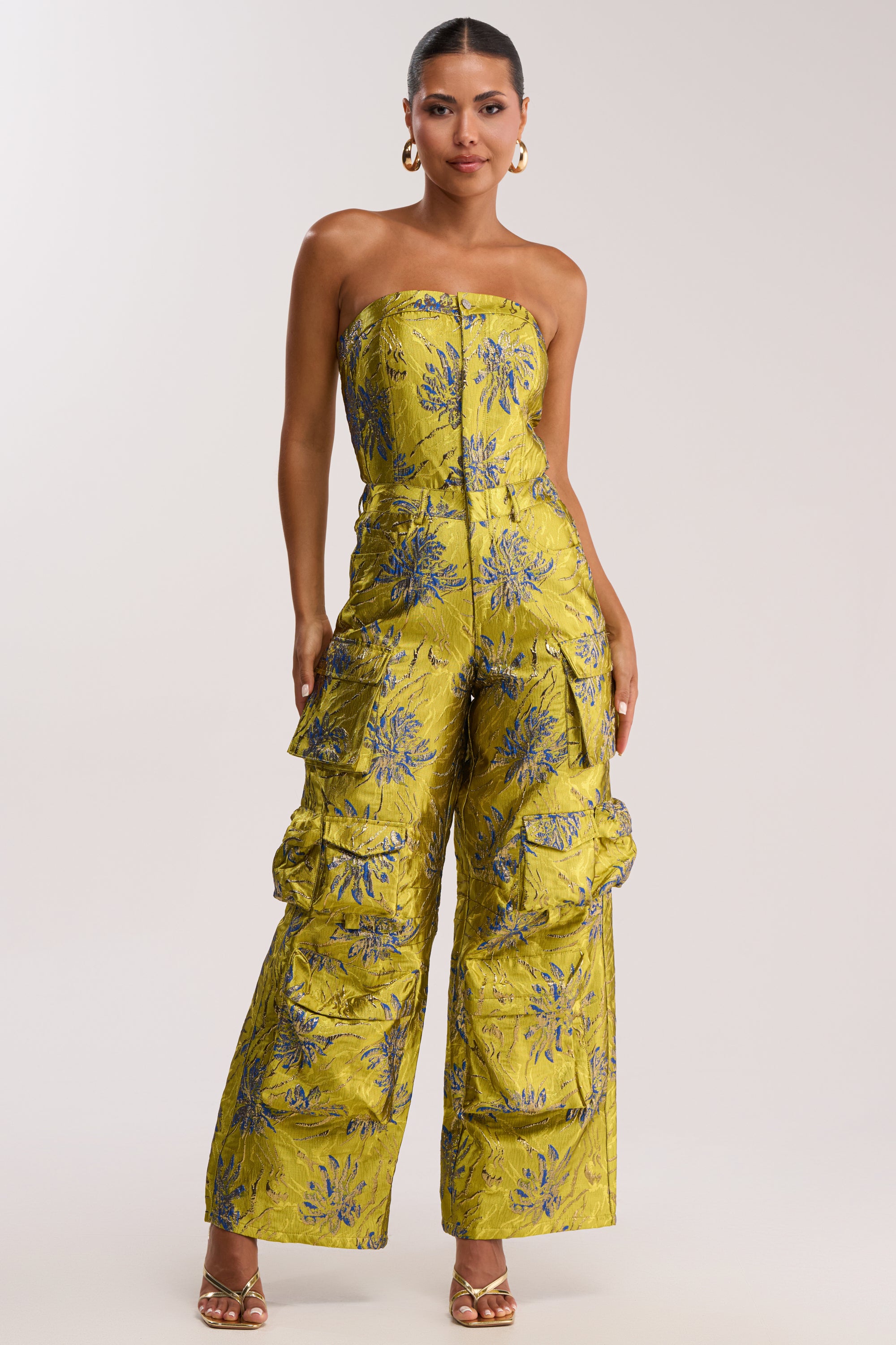 A woman stands against a plain light background wearing the NIKKI BROCADE JUMPSUIT IN GREEN, a strapless floral wide-leg jumpsuit with cargo pockets, styled with gold heels and hoop earrings.